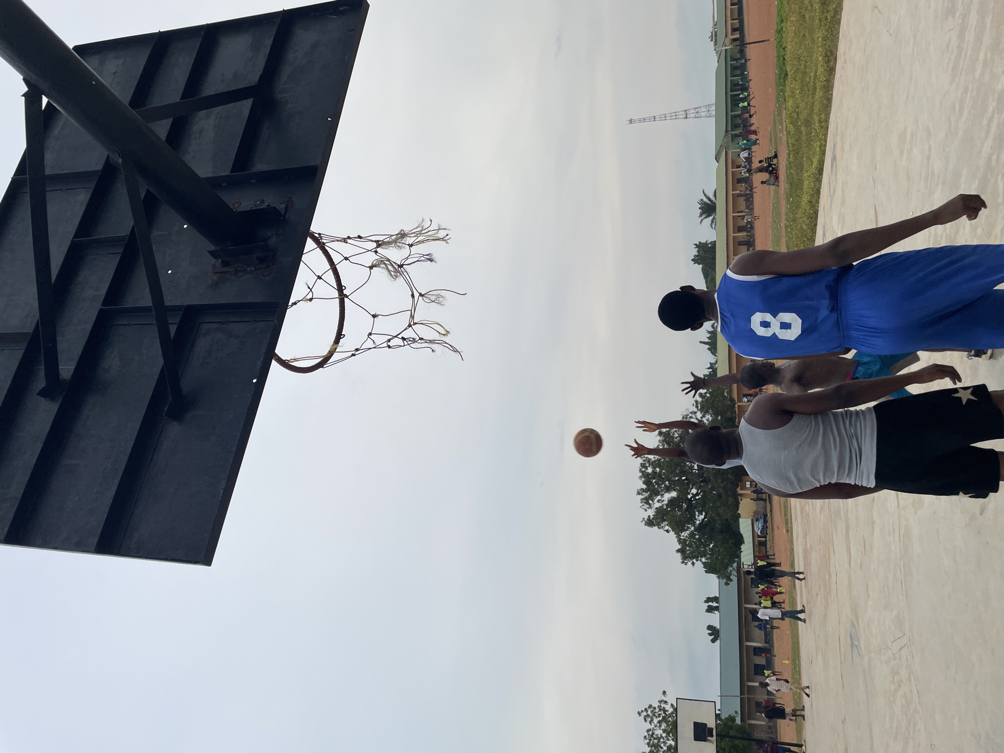 In Akwanga, Nigeria, on an outdoor basketball court, players are engaged in a game under an overcast sky. A basketball is captured mid-air, seemingly having just been released or in the process of being contested. Three young men are prominently featured in the foreground, two of whom are actively reaching for the ball, their arms extended upwards. One of these players is wearing a grey tank top and black shorts, while the other is in a blue basketball jersey with the number '8' on the back. A third individual, partially visible, stands to the left. In the background, other people are scattered across the court and on a reddish-brown field adjacent to the court. Several low-lying buildings with light-colored walls and green roofs are visible, suggesting a school or community center. Trees dot the landscape, and a tall, lattice-like tower stands in the far distance. The court surface appears to be concrete.