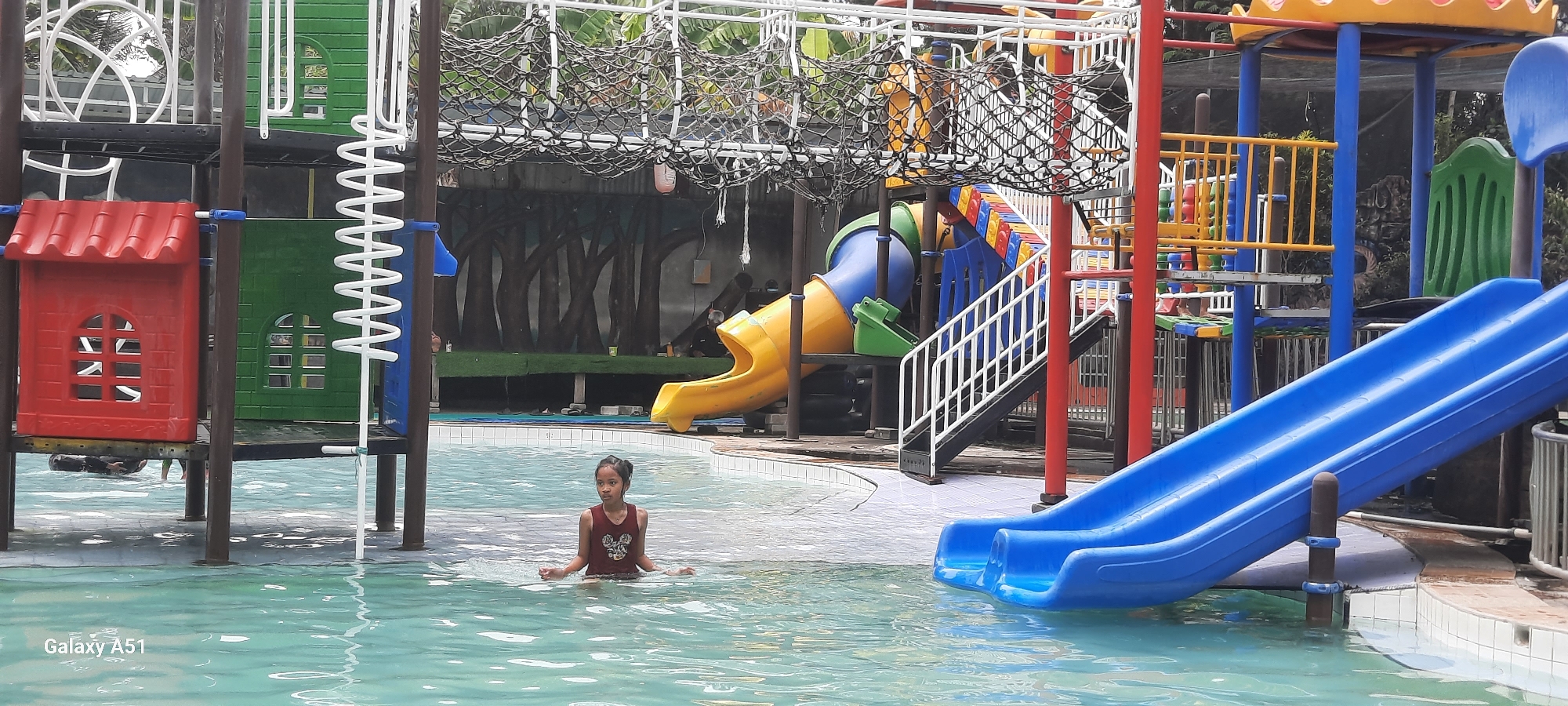 The image depicts a water park with a playground structure. In the foreground, a young girl is standing in shallow water, looking towards the camera. The playground features multiple slides, including a bright blue slide that extends into the water, a yellow slide, and a green tube slide. Various colorful elements, such as red and green playhouses and a climbing net, are visible. The background shows a mural of trees and foliage. The water is clear and reflects the surrounding structures. The water park appears to be located in a tropical or subtropical environment, as suggested by the lush vegetation. The "Galaxy A51" watermark indicates the device used to capture the image, and the location is identified as Popoh, Indonesia.