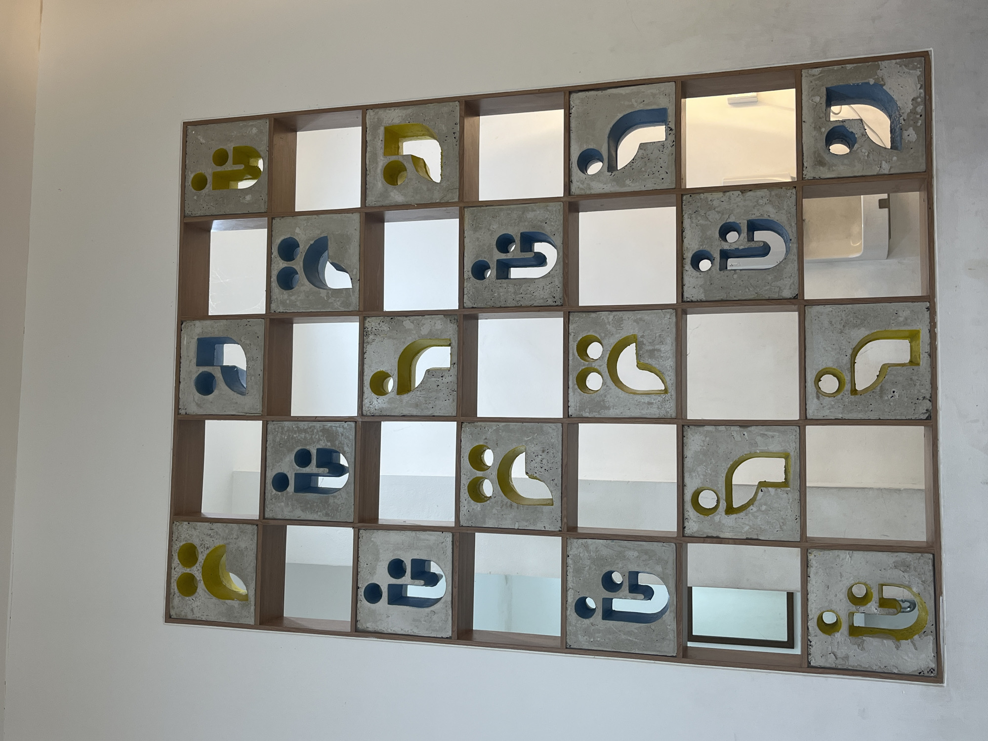 The image displays a grid of concrete tiles, each featuring an abstract cutout design. Some tiles have a yellow or blue colored interior within the cutout, while others are uncolored concrete. The tiles are arranged in five rows and five columns, with wooden dividers between them. This structure appears to be a wall or partition, with open spaces behind some of the tiles revealing a white wall and possibly doorways or alcoves. The overall aesthetic suggests a modern interior design. The location is identified as Tangerang, Indonesia. There are no people or animals visible in the image.