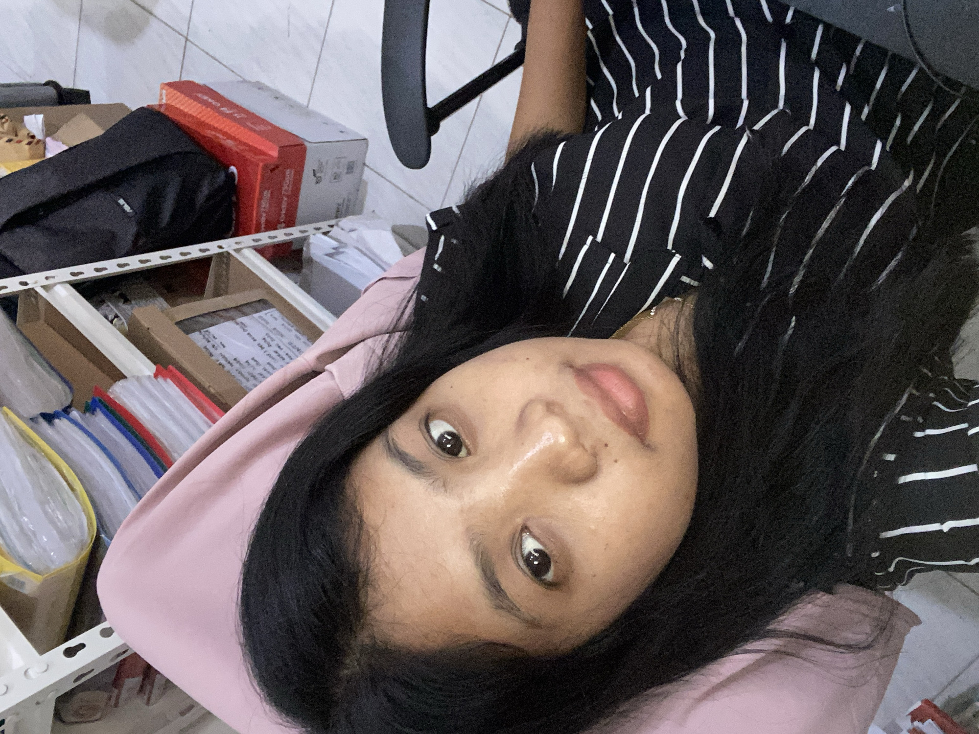 A person with dark hair and light skin is lying down, facing the camera. They are wearing a black and white striped shirt. Behind them is a light pink surface. To the left, there is a shelf with various objects including a black bag, red and white boxes, folders, and other items. The background suggests an indoor location, possibly an office or storage area. The picture may have been taken indoors, possibly in Negara, Indonesia based on the user's request.