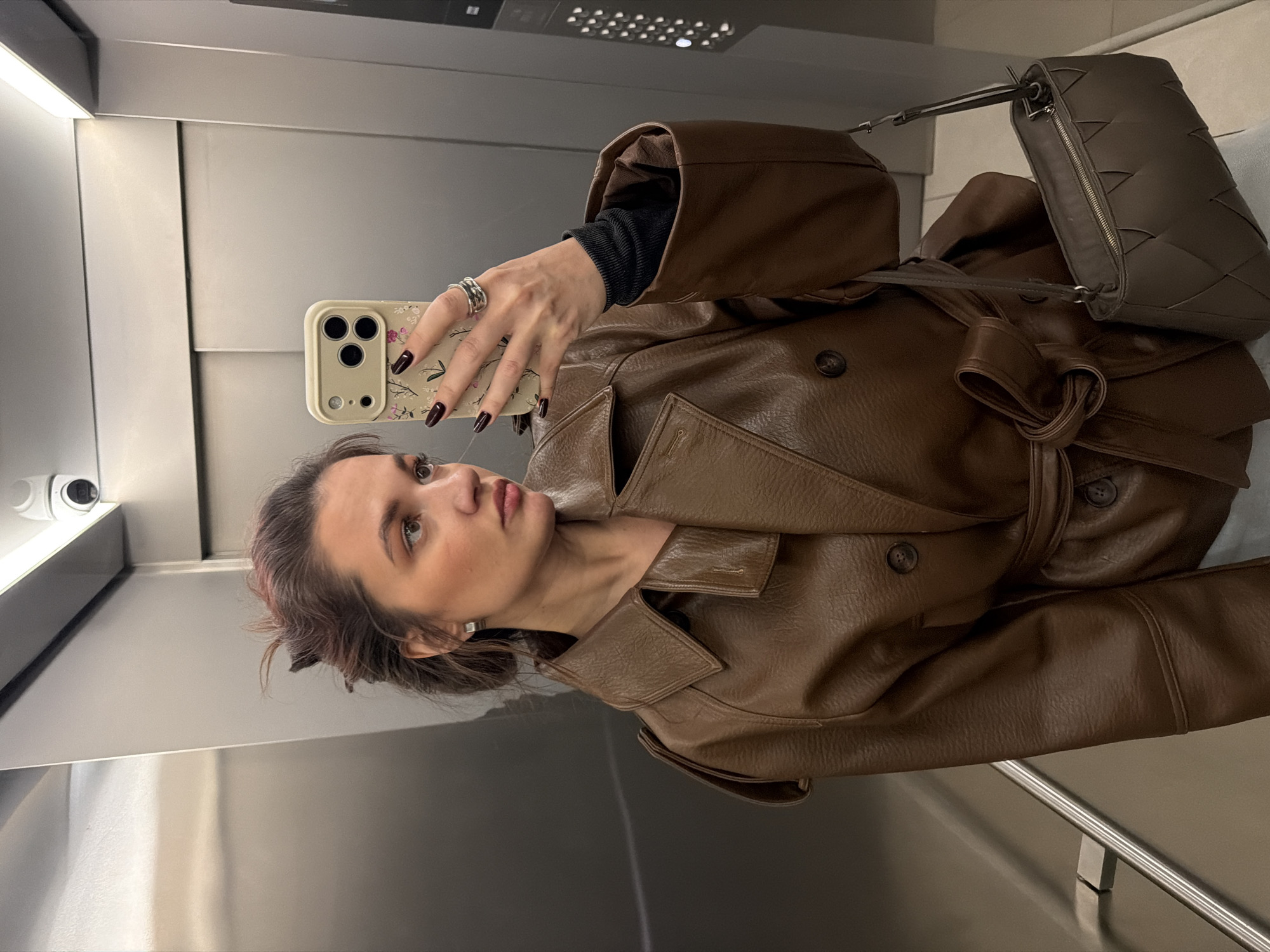 A young adult female is depicted taking a selfie within an elevator in Belgrade, Serbia. The precise location within Belgrade is not visually identifiable. The female, positioned slightly left of center, holds a light-colored smartphone with a floral-patterned case in her right hand, capturing her reflection. She has brown hair styled in an updo and wears a brown faux-leather double-breasted trench coat with a belted waist over a black long-sleeved top. She wears square silver earrings and a silver ring on her right ring finger; her fingernails are painted dark maroon. A dark brown woven shoulder bag with an open top hangs from her right shoulder. The elevator interior features brushed metallic walls, a horizontal ceiling light fixture, and a white dome-shaped security camera mounted on the ceiling in the upper right. A metallic handrail is visible on the left, and a black elevator control panel with illuminated buttons is visible on the right.
