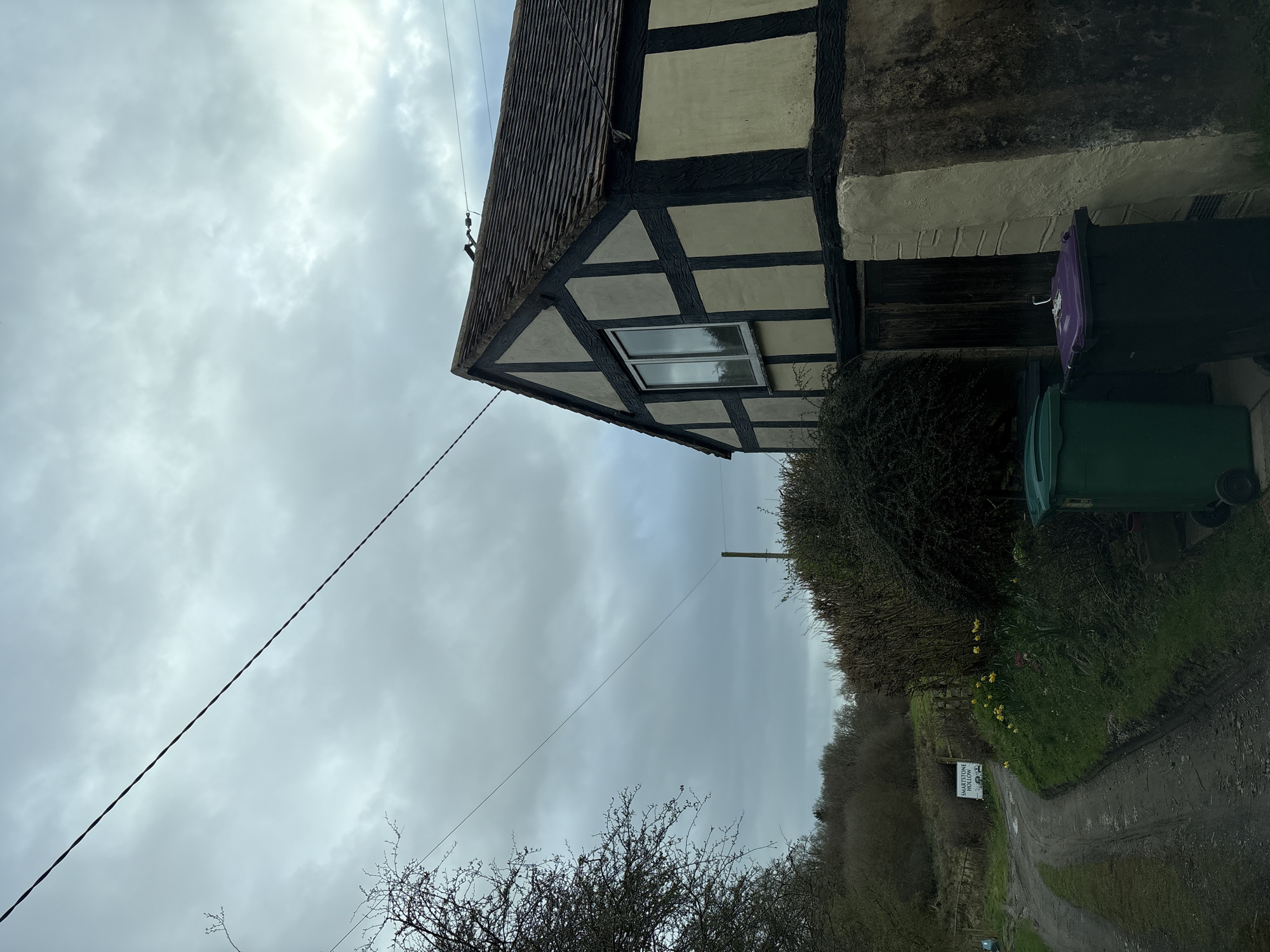The image depicts the exterior of a half-timbered house in Shelve, United Kingdom, under a cloudy sky. The house has a distinctive architectural style with dark timber framing contrasting against lighter infill panels. A single window is visible on the side of the house. Two wheelie bins, one purple and one green, are positioned adjacent to the house. A hedge is growing near the house. The scene includes a dirt path and a sign reading "SMARTSTONE HOLLOW". A power line stretches across the sky. Bare trees and bushes are visible in the background.