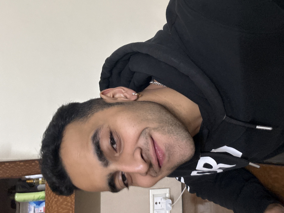 A young man with short dark hair and an earring poses in a black hoodie. He has fair skin, dark eyes, and light facial hair. A silver chain is visible at his neck. The background is a beige wall with a light switch and some household items on a wooden surface.