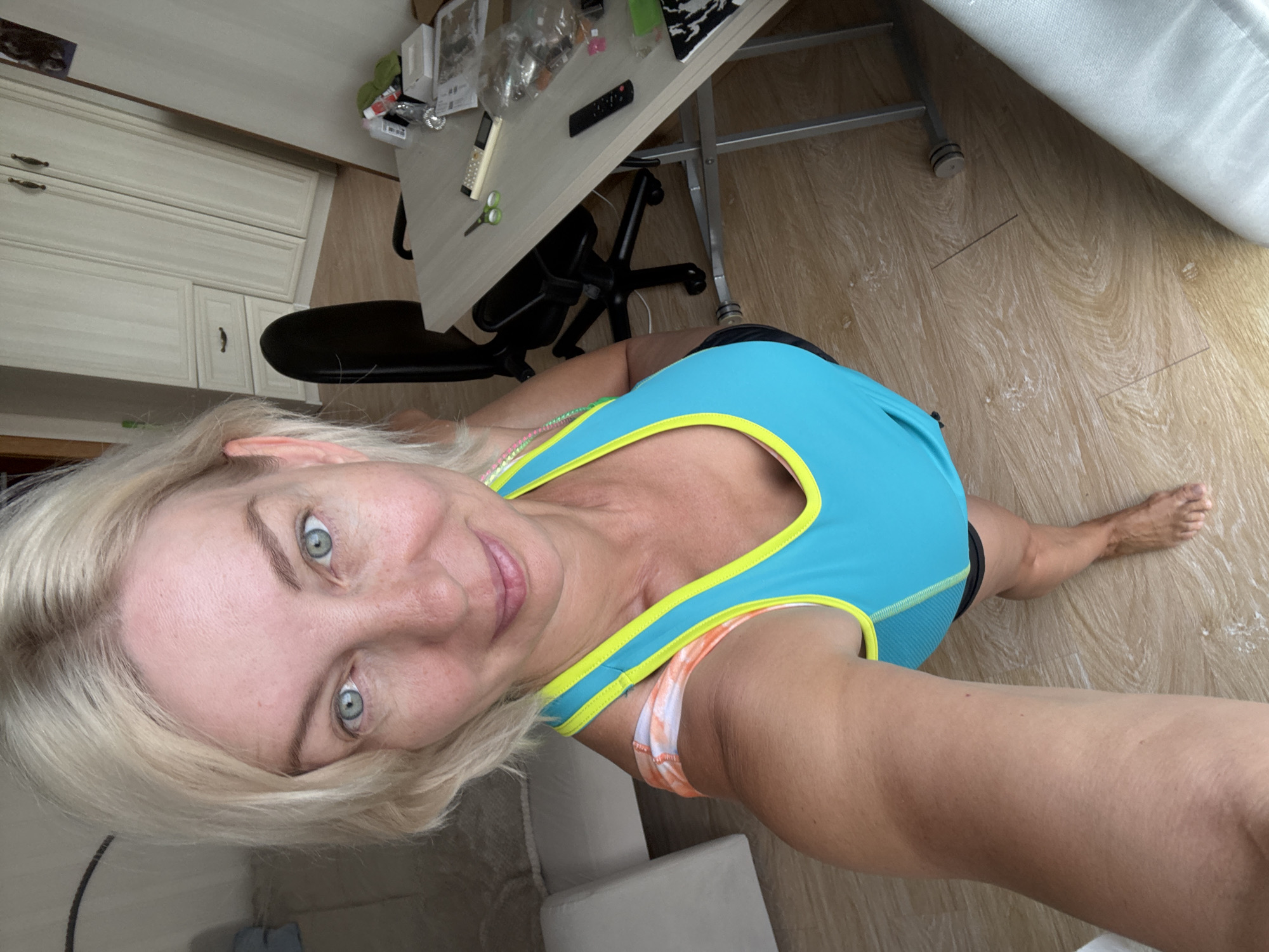 A woman with blonde hair and blue eyes, wearing a blue and yellow tank top and black shorts, stands in a room with light wood floors. She is holding a phone in her right hand, taking a selfie. A desk with a computer monitor, office chair, and various items is visible behind her, along with a tall cabinet with drawers and doors. The overall impression is that of an indoor personal space, possibly a home or office, in Sochi, Russia.