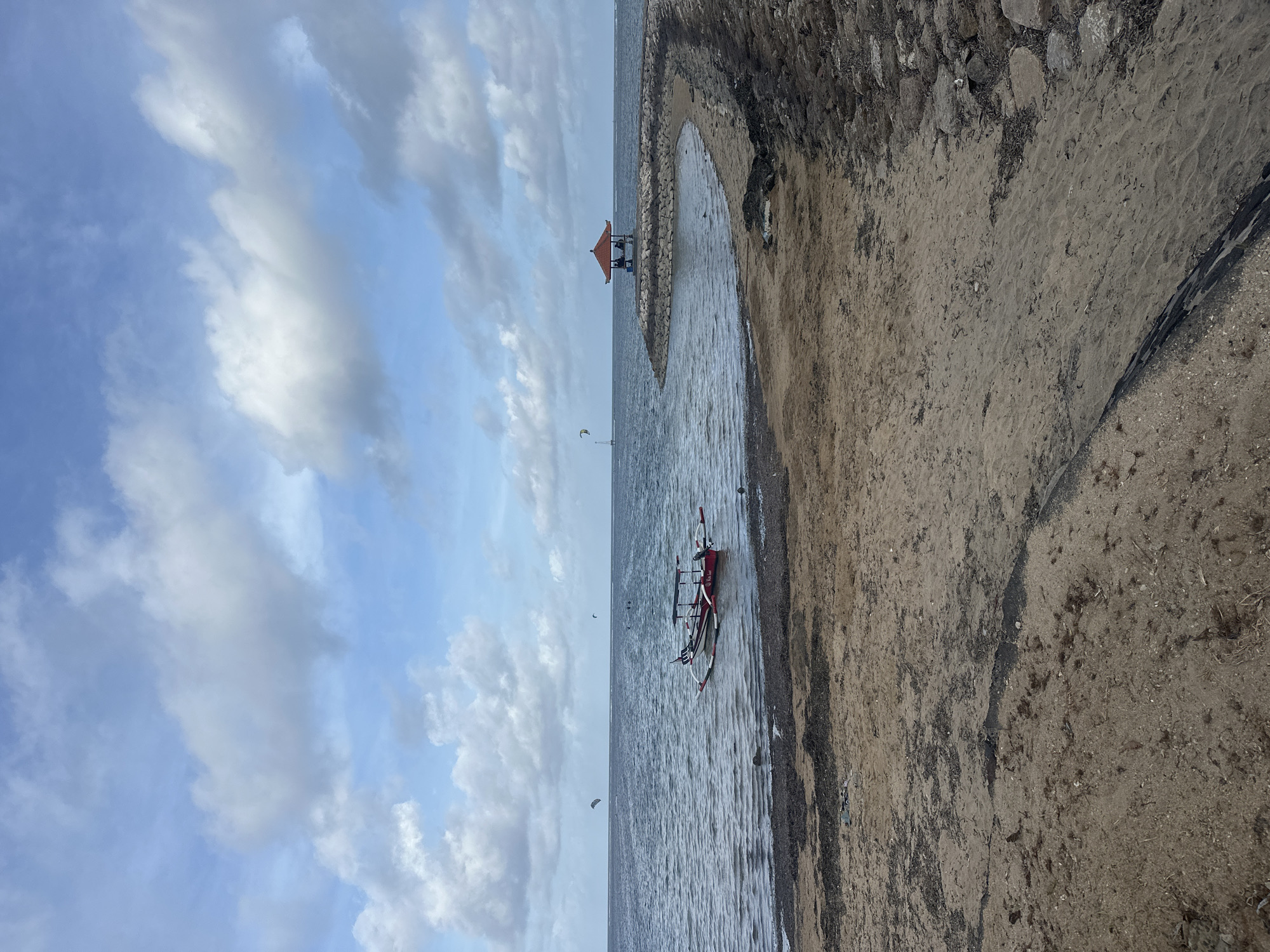 The image presents a coastal scene in Denpasar, Indonesia. The composition is split between the sky on the left, filled with cloudy blue hues, and the beach on the right. In the center is the water's edge. A traditional Indonesian outrigger boat is beached at the shoreline. Further out in the water, two kite surfers are visible. A small structure, likely a gazebo, stands on the horizon. The beach consists of sand and a low retaining wall made of rocks. The overall scene portrays a calm, coastal environment.