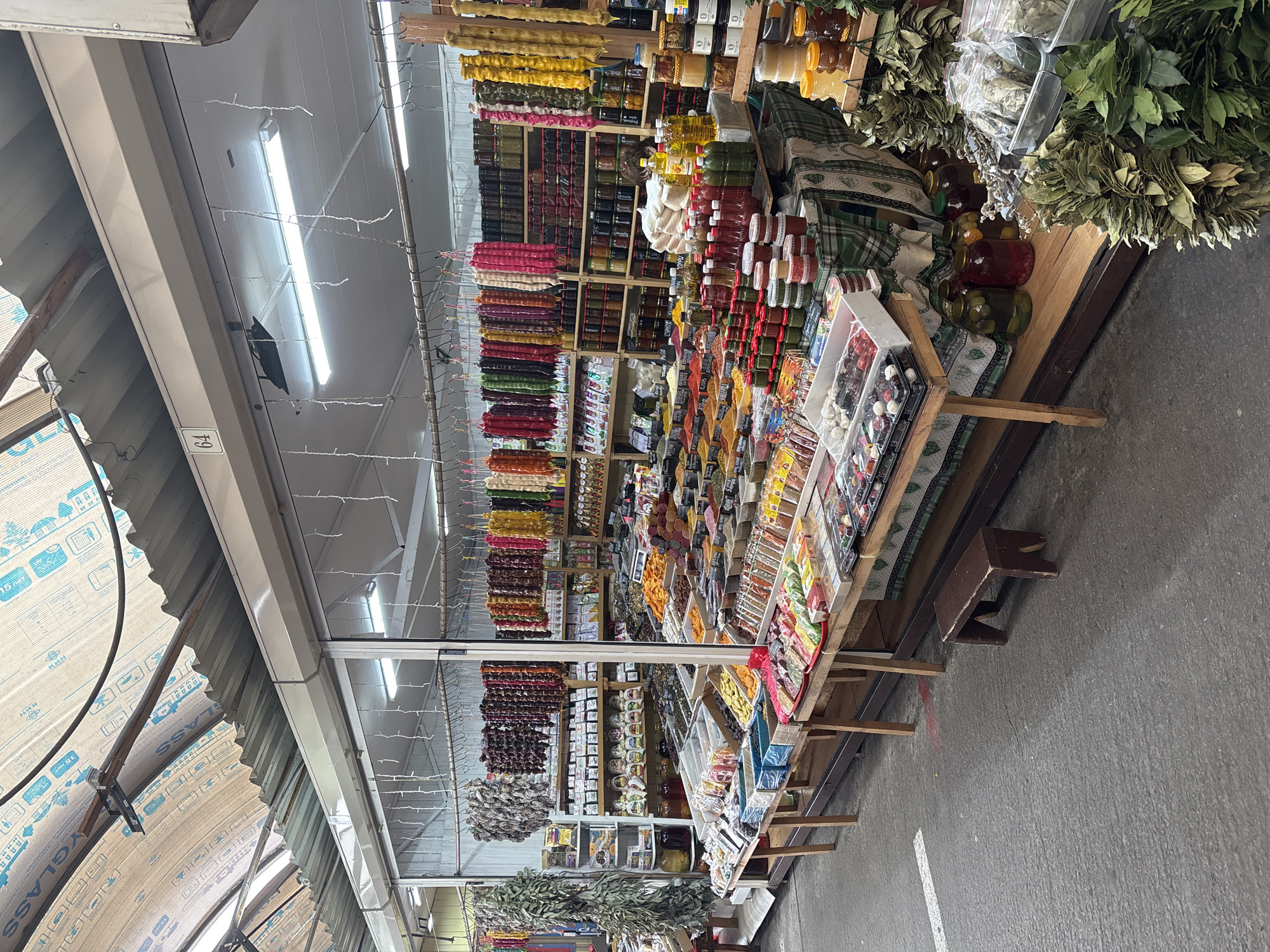 The image depicts a market stall in Sochi, Russia, filled with a variety of goods. Numerous strings of colorful, sausage-shaped sweets hang from the ceiling. Shelves behind the sweets are stocked with jars, packages, and containers holding various foods. The stall has a slanted display area featuring small boxes of sweets and spices. Bundles of bay leaves are positioned at the front edge of the stall. Overhead lighting fixtures are visible. The flooring is paved.