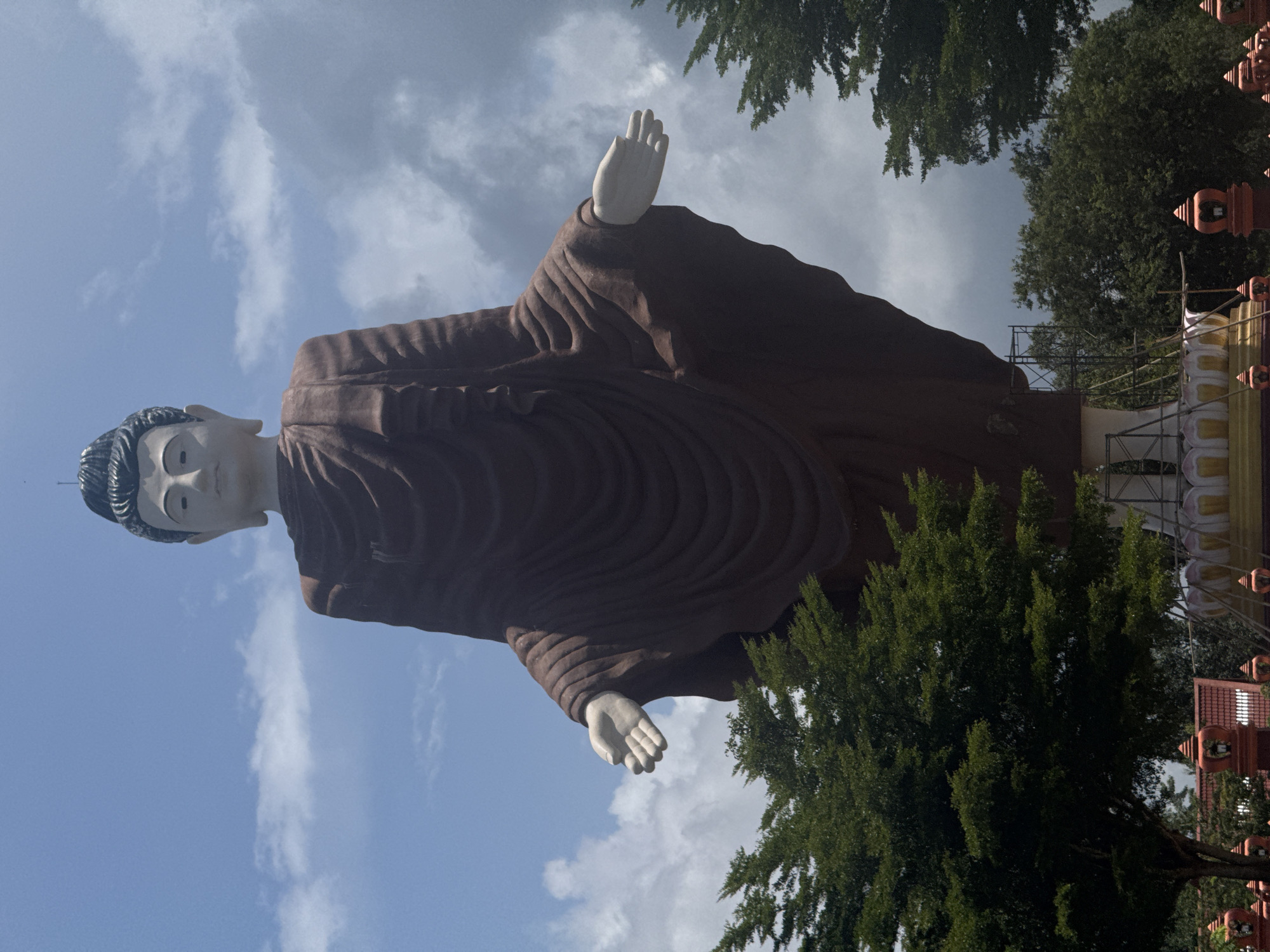A low-angle, full-shot captures a monumental, standing Buddha statue against a partly cloudy sky in Na Nuea, Thailand. The statue, draped in a dark brown robe, has its arms extended outwards, palms facing forward. The face of the Buddha is rendered in a pale, serene expression. In the foreground, lush green trees partially obscure the base of the statue, which appears to be undergoing construction or renovation, indicated by scaffolding and supporting structures. Decorative red and orange architectural elements are visible at the very bottom of the frame. The overall impression is one of grandeur and spiritual significance.