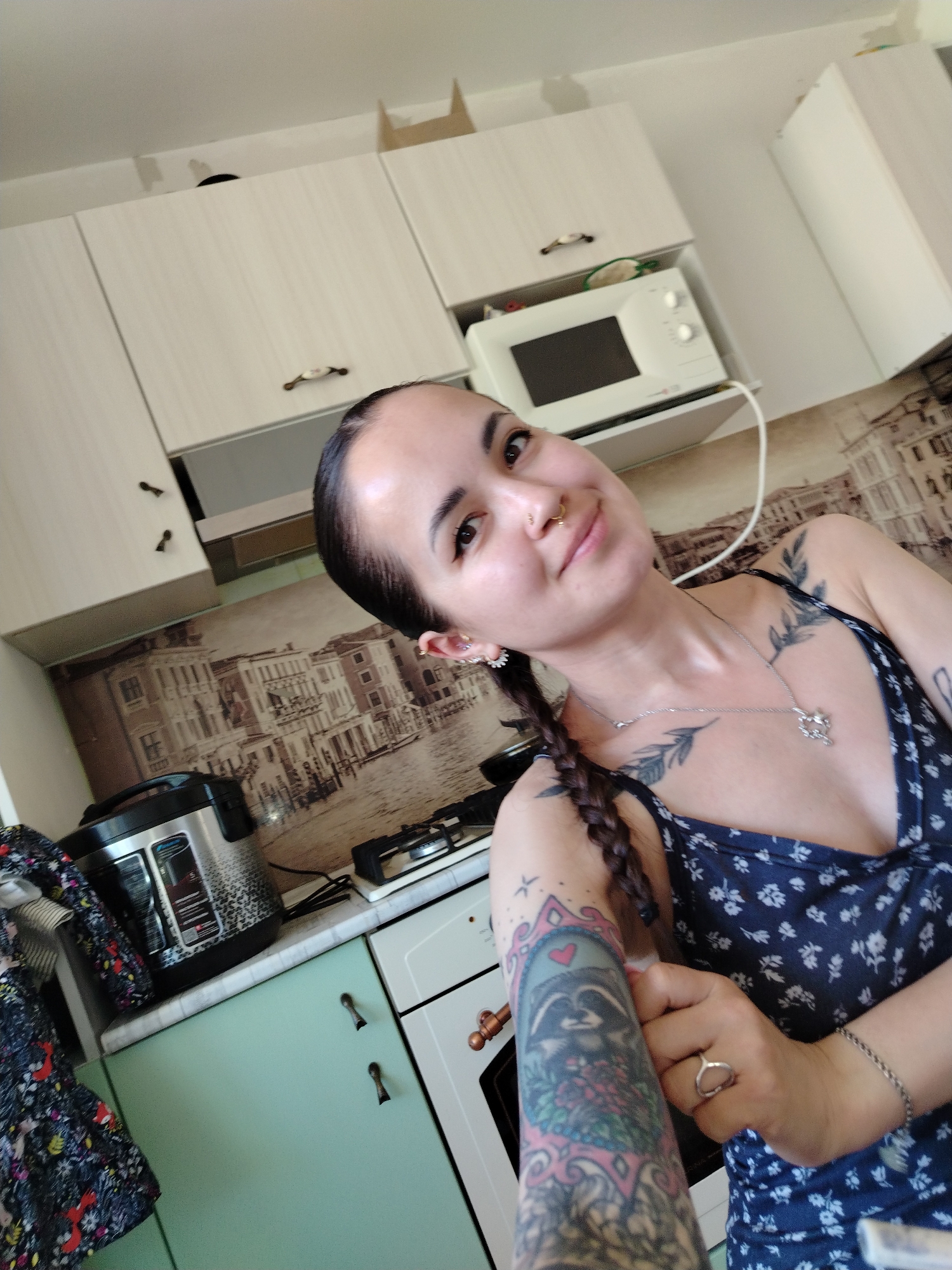 A person with a nose piercing, tattoos on their chest and left arm, and a braid is standing in a kitchen. The kitchen has white cabinets, a microwave, a stovetop, and a pressure cooker. The backsplash has a printed image of buildings along a canal. The location appears to be in Noviy, Russia, based on the background image.