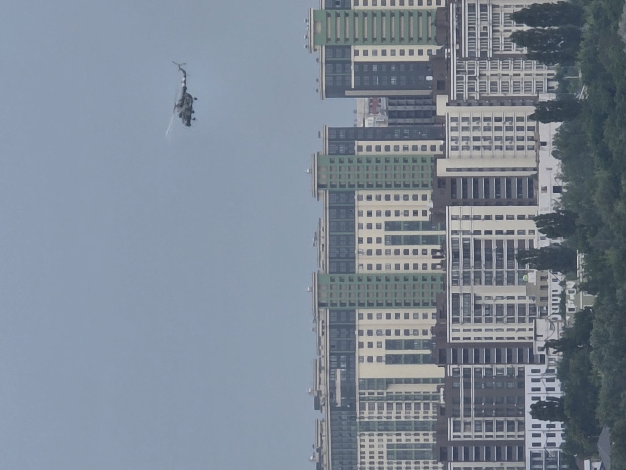 The image shows a military helicopter flying over a cityscape. The helicopter is a dark color and appears to be a combat helicopter. It is flying at a high altitude, and the rotor blades are blurred, indicating movement. In the background, there are several tall apartment buildings. The buildings are modern and appear to be residential. They are multi-story structures with many windows. The sky is overcast, and the overall lighting is muted. The image appears to have been taken from a distance, possibly from another building or a drone. The scene is likely an urban environment, possibly in Kyiv, Ukraine, given the context of a military helicopter in flight. There are no people visible in the image. The environment is characterized by high-rise architecture and an aircraft in the sky.