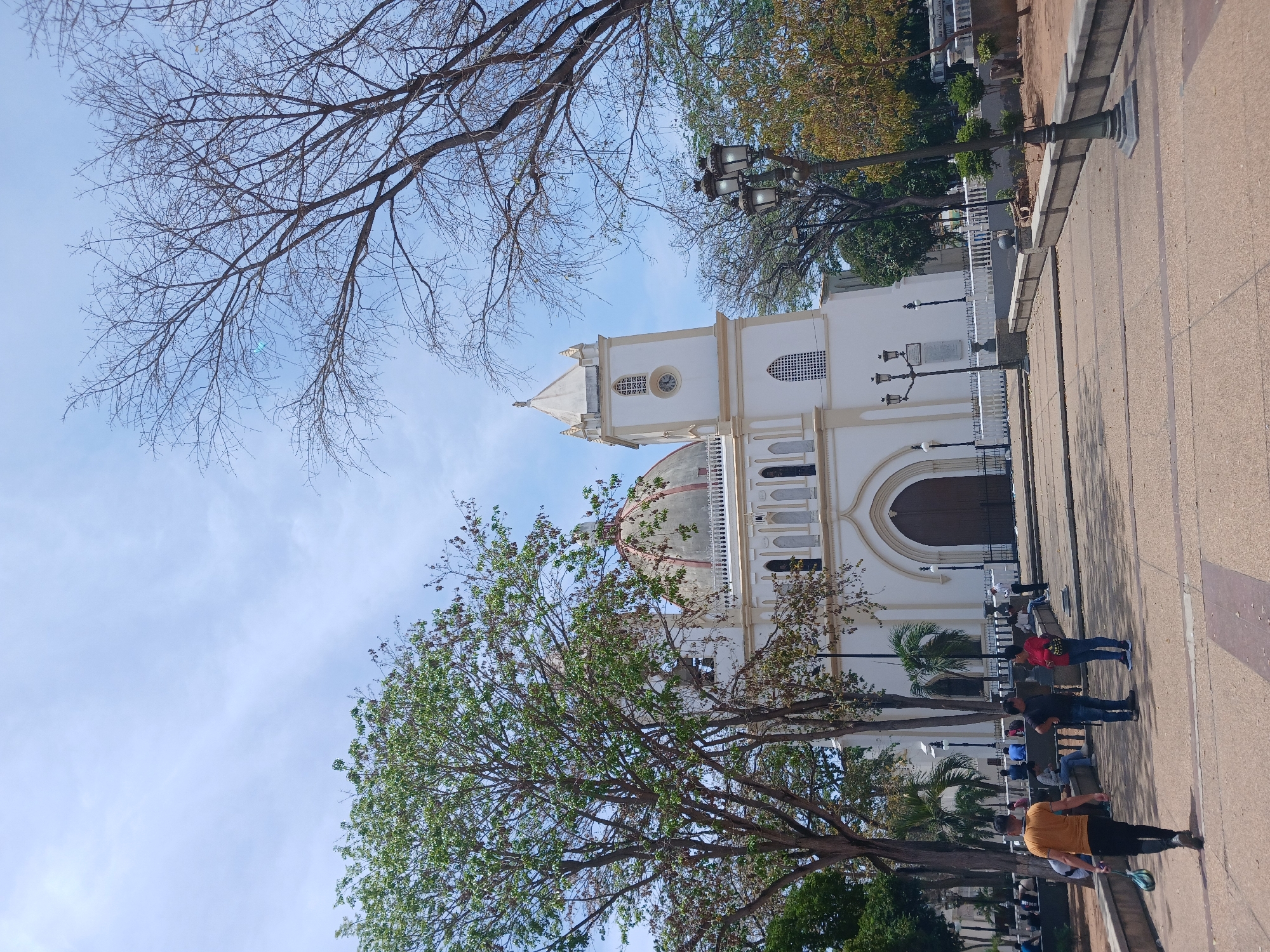 The image depicts the Basilica Menor de Nuestra Señora del Valle, a church in Porlamar, Venezuela. The white building features a domed roof and a bell tower, partially obscured by trees with green and bare branches. A paved walkway runs along the side of the church, with several people sitting on benches, some wearing casual clothing. Lampposts and a white fence are visible along the walkway. The sky is blue with some clouds.