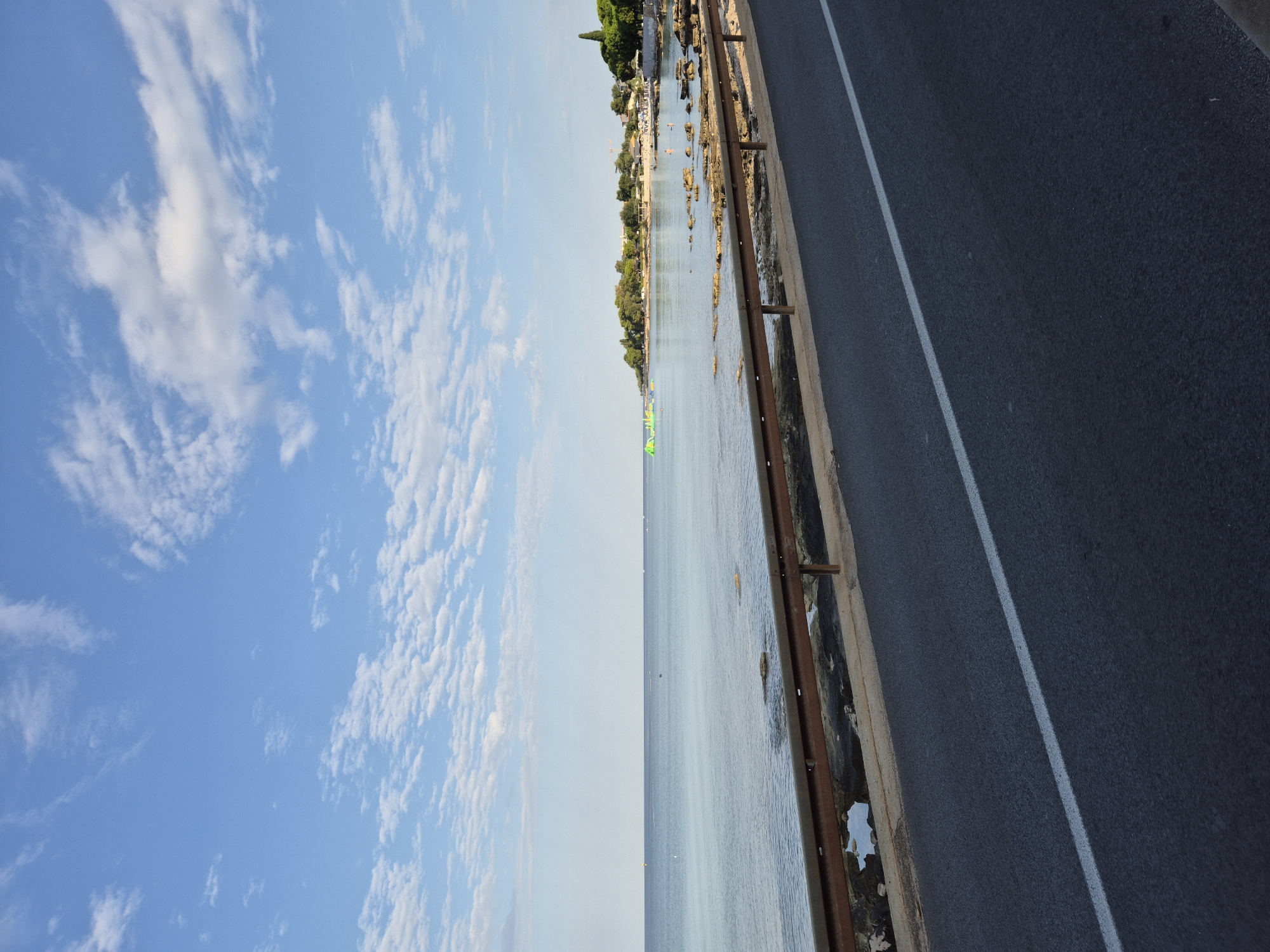 The image shows a coastal road in Zambratija, Croatia, with the Adriatic Sea on the left and a tree-lined shore on the right. The road is paved and has a white line marking the lane. A rusty metal guardrail separates the road from the rocky shoreline. The sea is calm and appears shallow near the shore, with some rocks visible. In the distance, on the water, there are inflatable water attractions, and people can be seen on a sandy beach. The sky is blue with scattered white clouds. The overall scene depicts a typical summer day at a coastal resort.