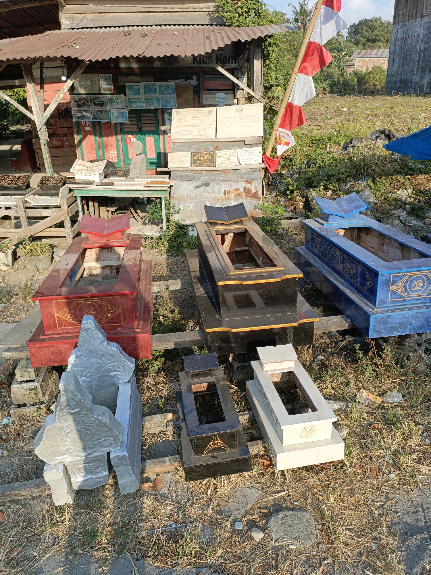 In the city of Bulukumba, Indonesia, on an outdoor lot, an array of finished and unfinished grave markers are displayed. A small, rustic stall with a corrugated metal roof and red and green painted walls stands in the background, displaying various religious texts and framed images. In front of the stall, a wooden table is laden with more grave markers, some adorned with decorative lettering. Several complete graves, including a red one with gold accents, a black one with gold trim, and a blue one with intricate gold patterns, are arranged on wooden planks. Scattered around these are smaller grave markers and a large, rough-hewn grey stone structure. The ground is covered with dry grass and debris, and a red and white Indonesian flag flutters from a bamboo pole. The scene suggests a small business selling gravestones. No people are present in the image.