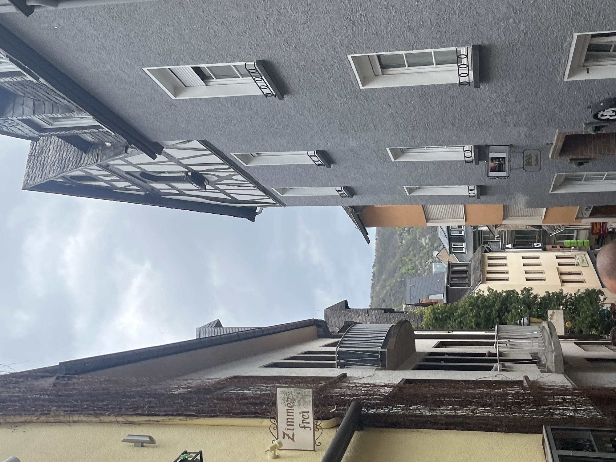 The image depicts a street scene in Cochem, Germany. Buildings line the narrow street, showcasing traditional German architecture with gabled roofs and half-timbered construction. A building with gray walls and white framed windows and security bars is visible at the top of the frame. On the bottom of the frame is a building that has the words "Zimmer frei" (Rooms Available). The sky is overcast. In the distance, more buildings and green hillsides are visible.