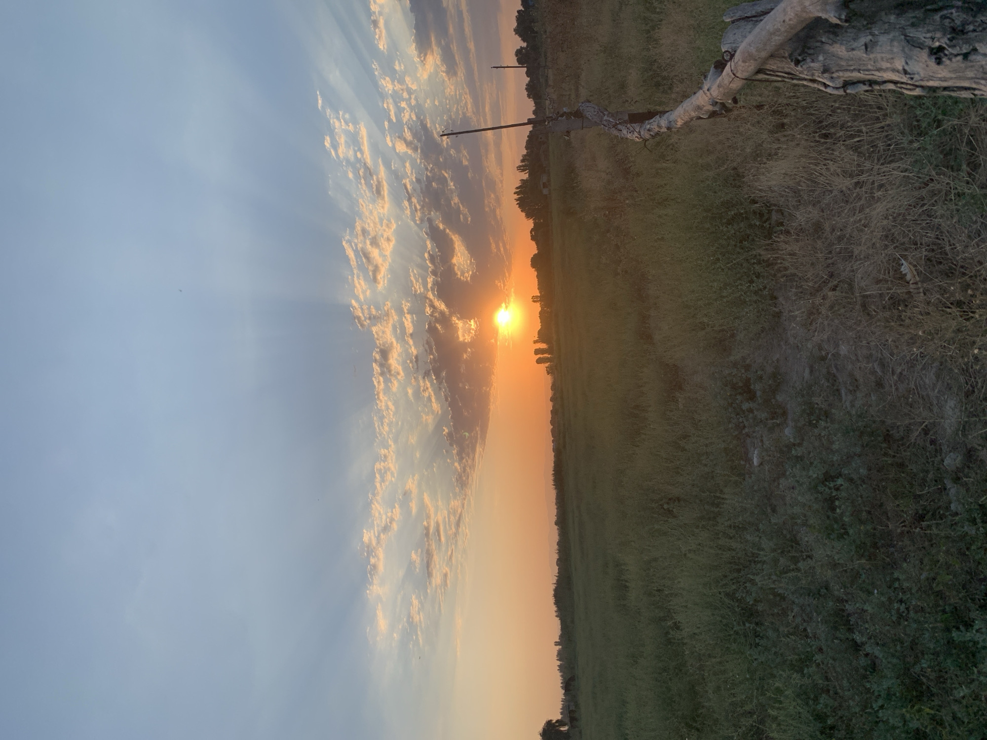 The image depicts a rural landscape at sunset. The sky is a gradient of blue and orange, with the sun visible on the horizon, casting light rays through scattered clouds. A field of green grass stretches across the foreground, leading to a line of trees and scattered structures in the distance. A weathered wooden fence runs along the right side of the frame, partially obstructing the view of the field. The location is identified as Komsomolskoe, Kyrgyzstan.