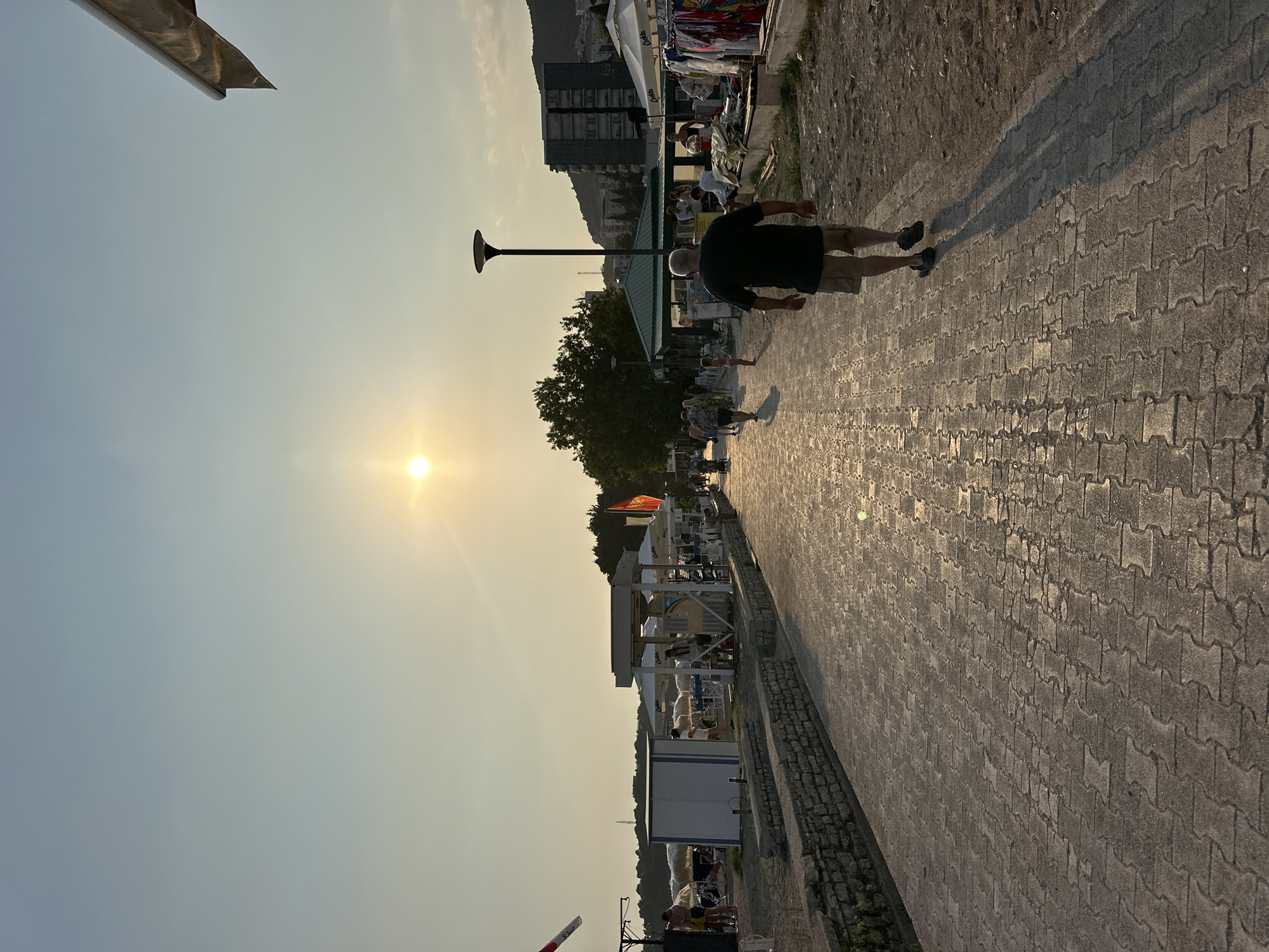 This is a horizontal, eye-level, medium-shot photograph taken in Boreti, Montenegro, during the golden hour. The sun is low in the sky, casting a warm glow and long shadows across the scene. The primary subject is a paved walkway made of interlocking rectangular stones, which reflects the sunlight.
On the right side of the walkway, a man in a dark t-shirt and khaki shorts is walking away from the camera, his shadow stretching out behind him. Further along the walkway, several other people are visible, some walking and some standing, suggesting a public promenade.
To the left of the walkway, there is a stone retaining wall. Beyond the wall are beach umbrellas and some small structures that appear to be shops or cafes. In the background, trees and buildings, including a multi-story structure, are visible, with hills or mountains behind them. A flag is visible on a pole to the right of the walkway. The sky is clear with a light haze. The overall atmosphere is that of a seaside town in the late afternoon or early evening.