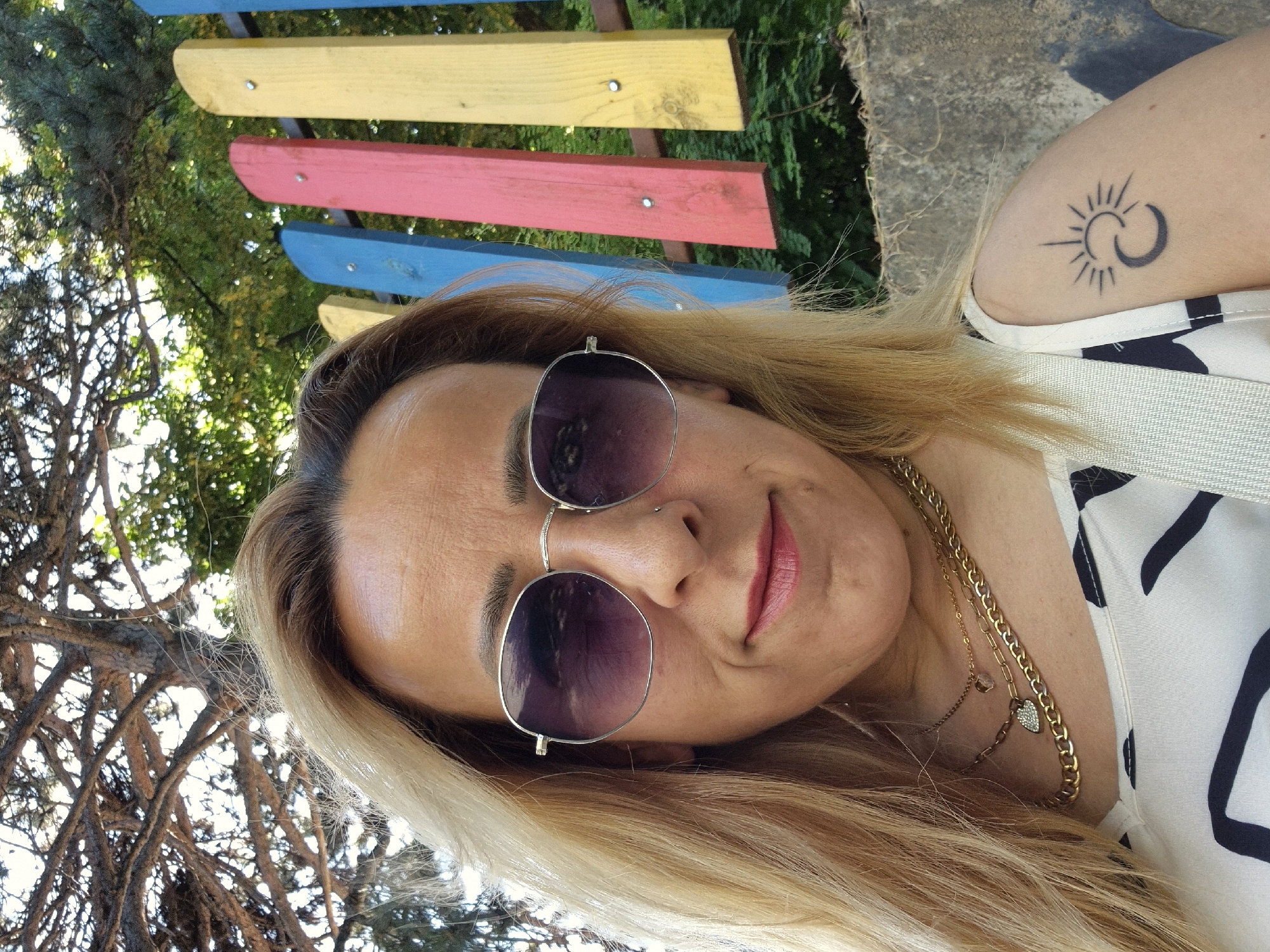 The image is a selfie of a woman with blonde hair, wearing sunglasses, a white and black patterned top, and multiple necklaces. She has a tattoo of a sun and moon on her left upper arm. Behind her is a colorful fence made of vertical wooden planks painted blue, pink, and yellow, and beyond that, greenery and tree branches are visible. A concrete wall is on the right side of the image. The setting appears to be outdoors.