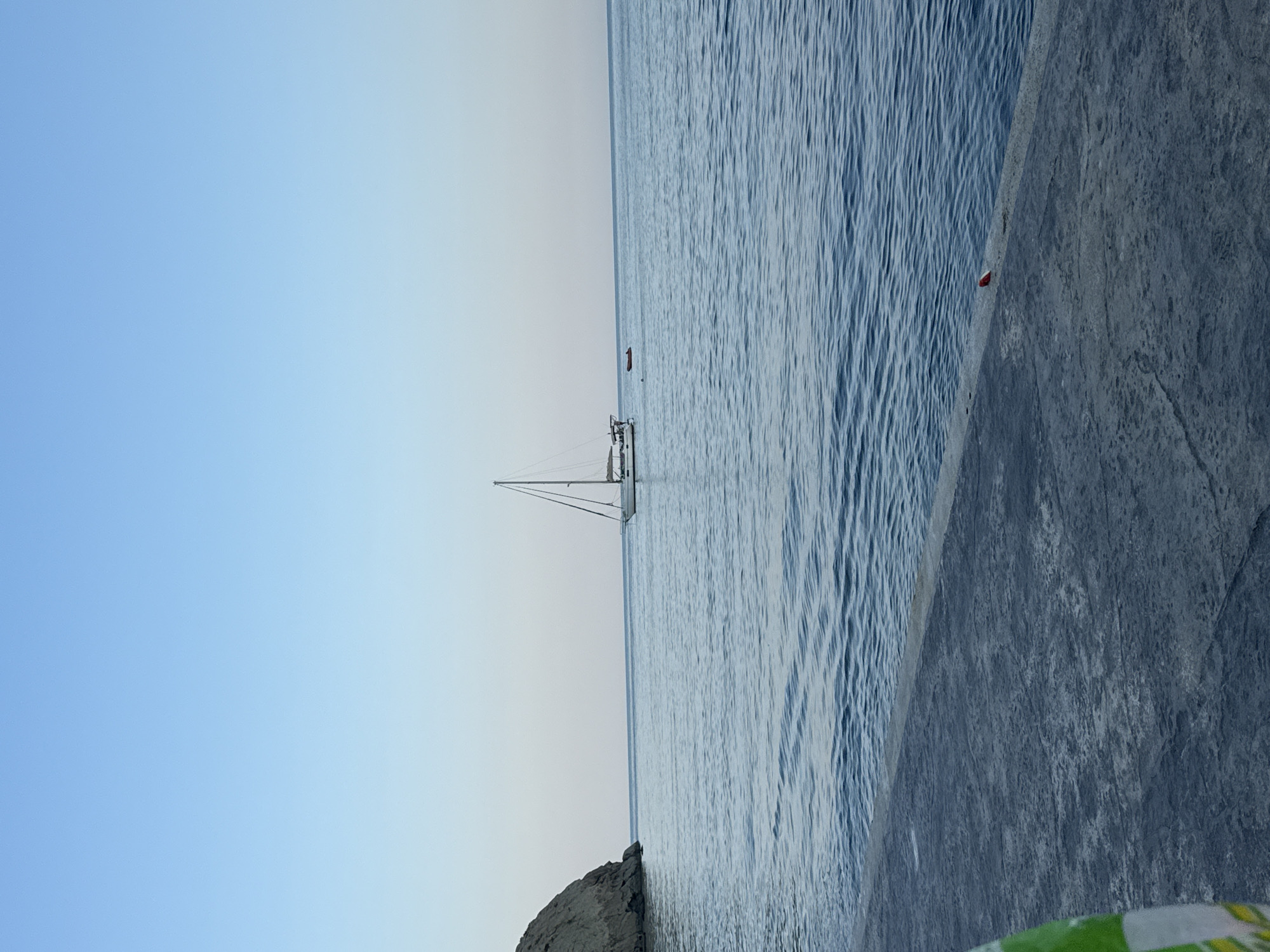 This is an eye-level, medium shot of a calm sea with a sailboat in the distance. The foreground is a concrete walkway. A rocky cliff is visible on the left side of the image. The sky is a clear, pale blue. There are no people or actions depicted in the image. The scene appears to be a coastal area.
