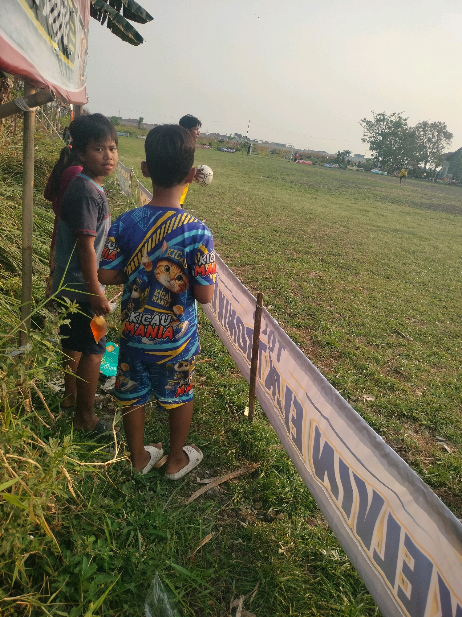 Two young boys are standing next to each other, looking away from the camera towards a grassy field. The boy on the left is wearing a dark t-shirt and shorts, and the boy on the right is wearing a brightly colored t-shirt and shorts with a cat graphic. A banner with text is stretched out in front of them, supported by wooden stakes. In the background, there are buildings and trees, suggesting an outdoor setting in a populated area, likely in Kalisari, Indonesia. The sky is overcast, and the sun appears to be setting.