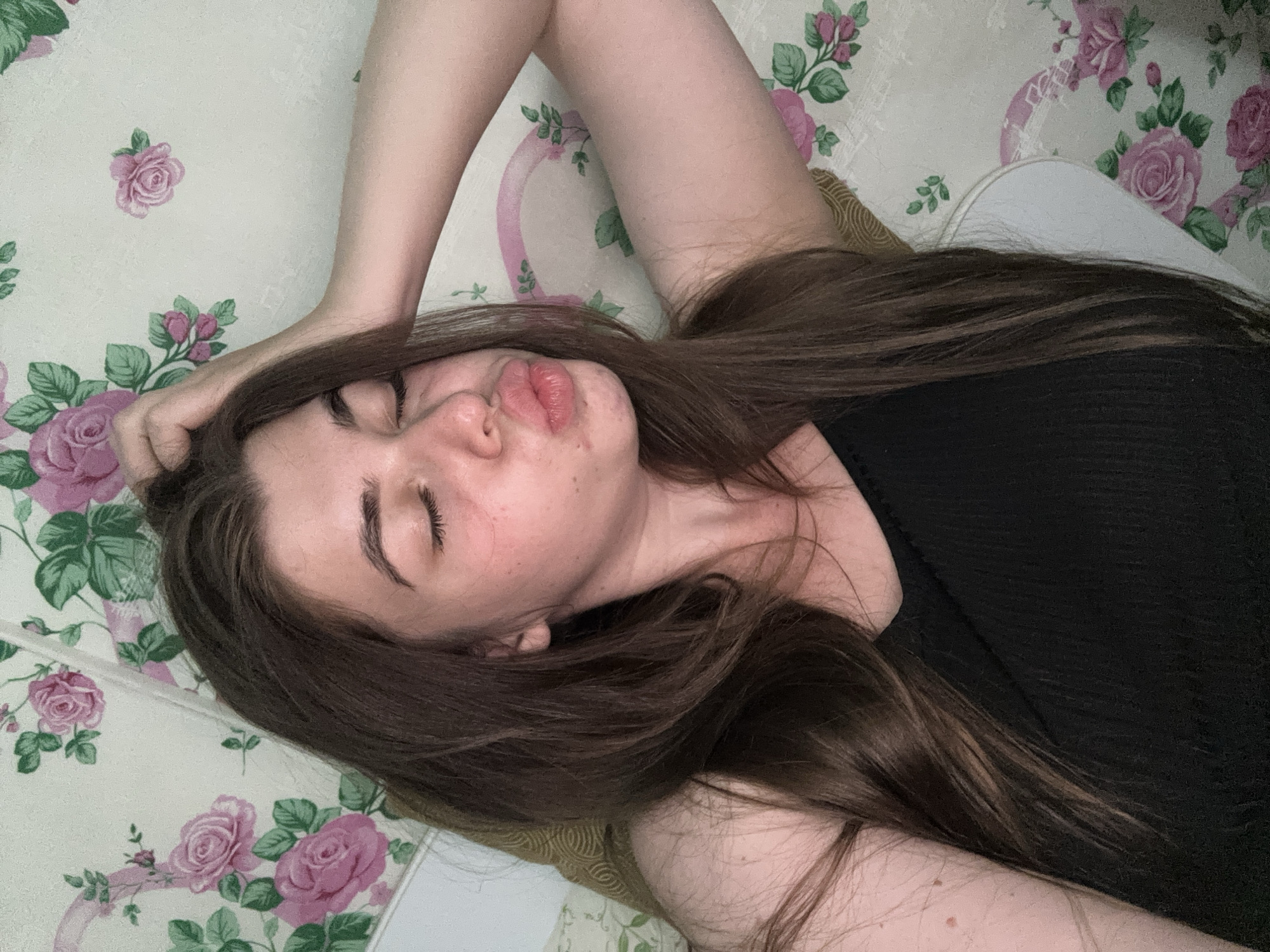 A young woman with long brown hair is lying down, with her right arm raised and hand on her head. Her eyes are closed, and her lips are pursed in a kiss shape. She is wearing a black tank top. The background is a floral wallpaper with pink roses and green leaves. The image appears to be taken indoors, possibly in a bedroom. There is no visible information within the image to suggest the location is Мещёры, Russia.
