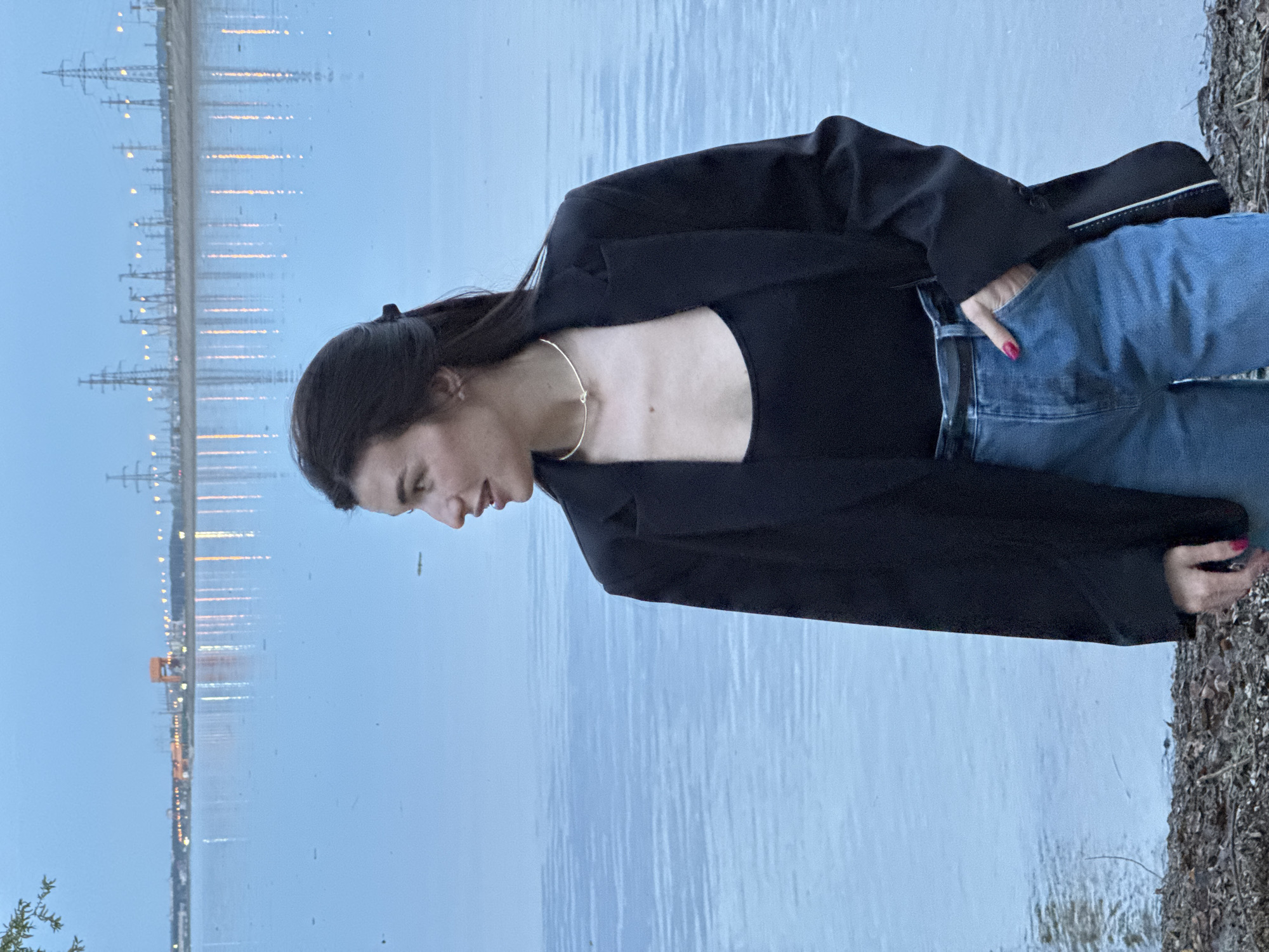 A woman stands by a body of water, likely the Volgograd reservoir in Noviy, Russia, given the presence of pylons reflected in the water, characteristic of the Volgograd Hydroelectric Station. The woman is wearing a black top and blazer, blue jeans, and a gold necklace. Her right hand is in her jeans pocket. The immediate environment consists of shoreline foliage.
