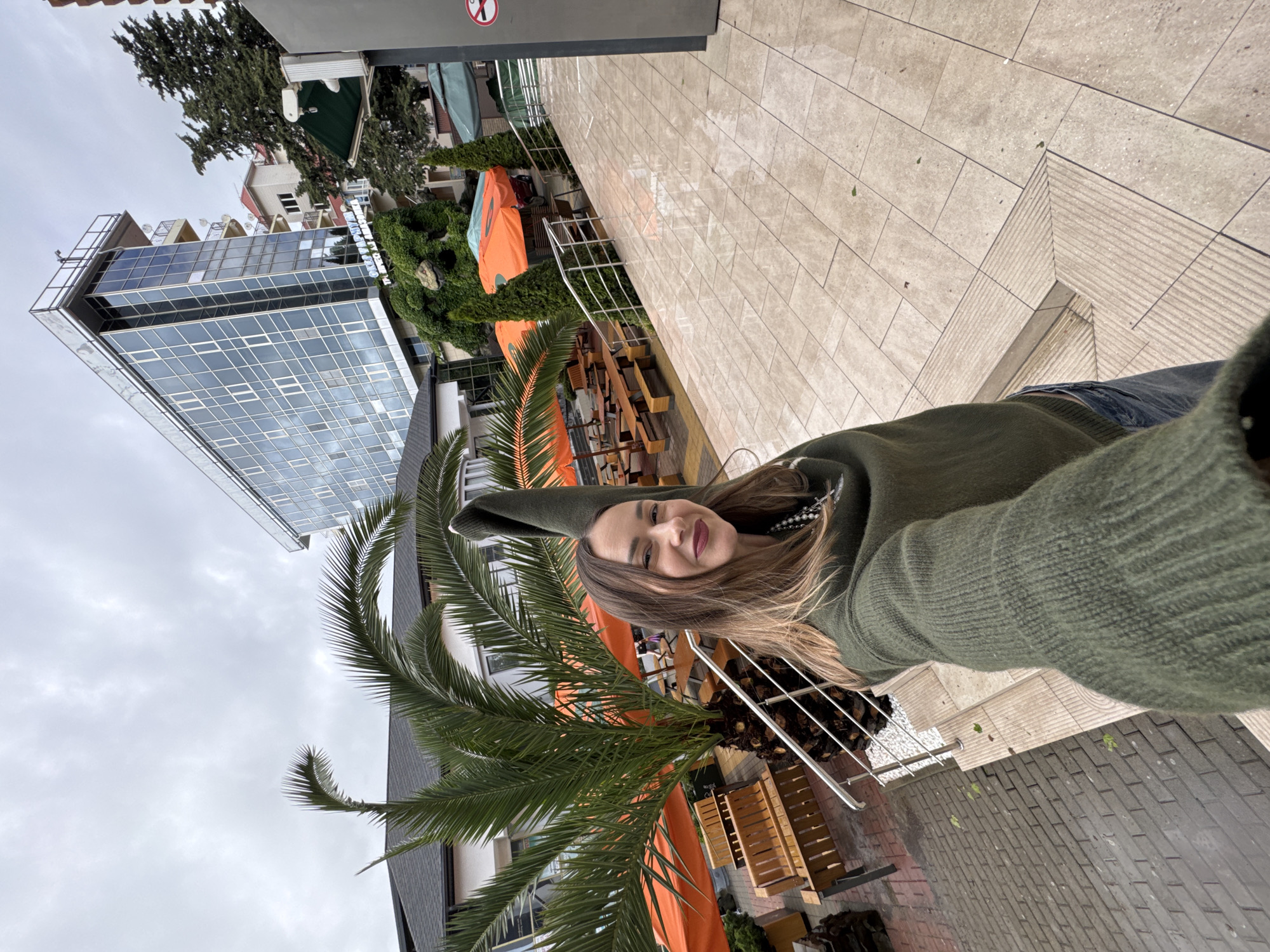 A woman is taking a selfie in Sochi, Russia. She is wearing a green hoodie and standing near a palm tree. Behind her is a modern building with glass windows and some cafes. The weather is overcast.