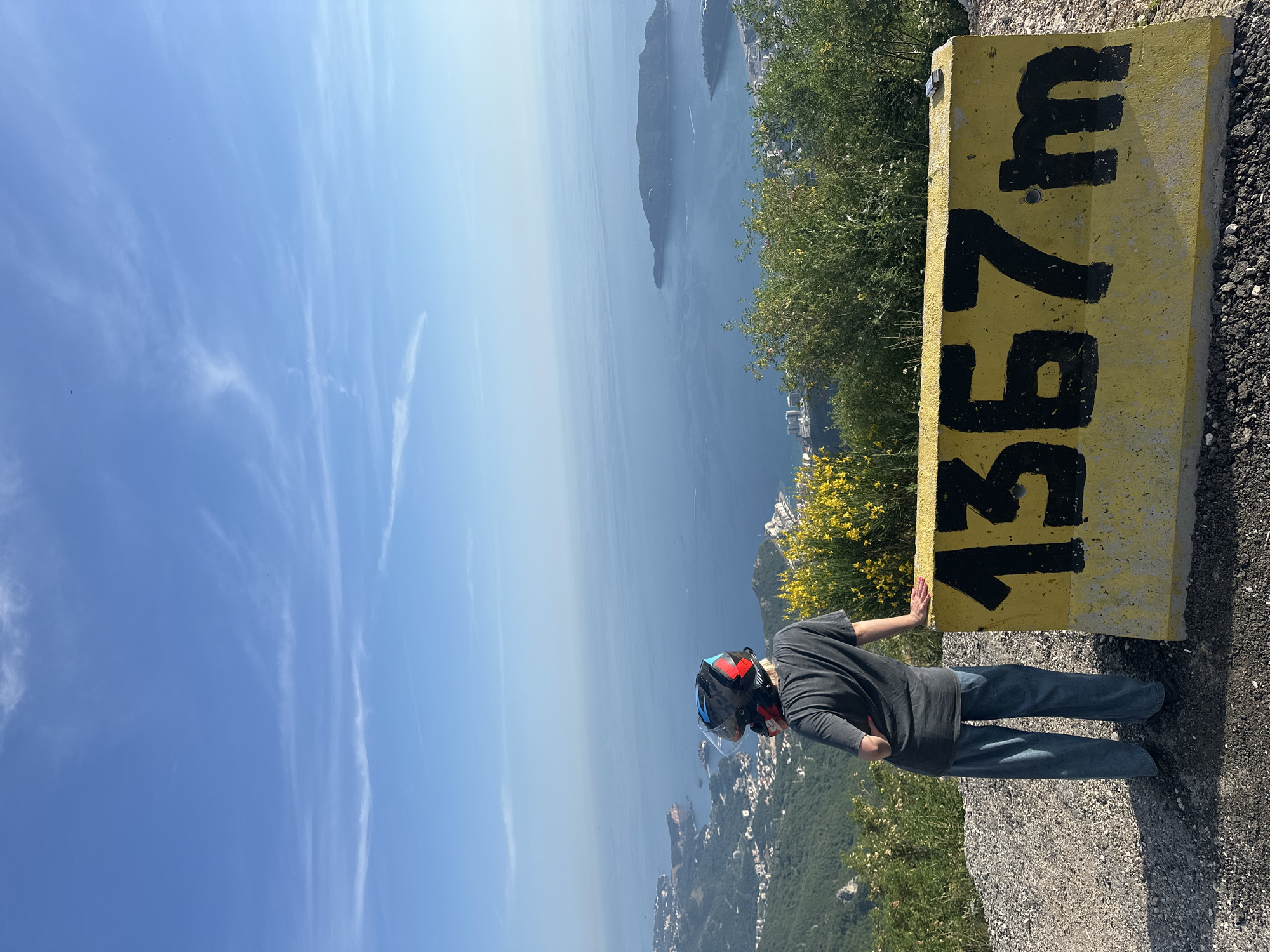 A medium shot depicts a person in casual clothing and a motorcycle helmet standing near a yellow concrete post marked "1367 m". The person is standing on a cliff overlooking the Adriatic Sea and part of the city of Brajici, Montenegro. The weather is sunny, with a clear blue sky dotted with wispy clouds. Green vegetation and yellow flowers grow near the post, adding to the scenic view.