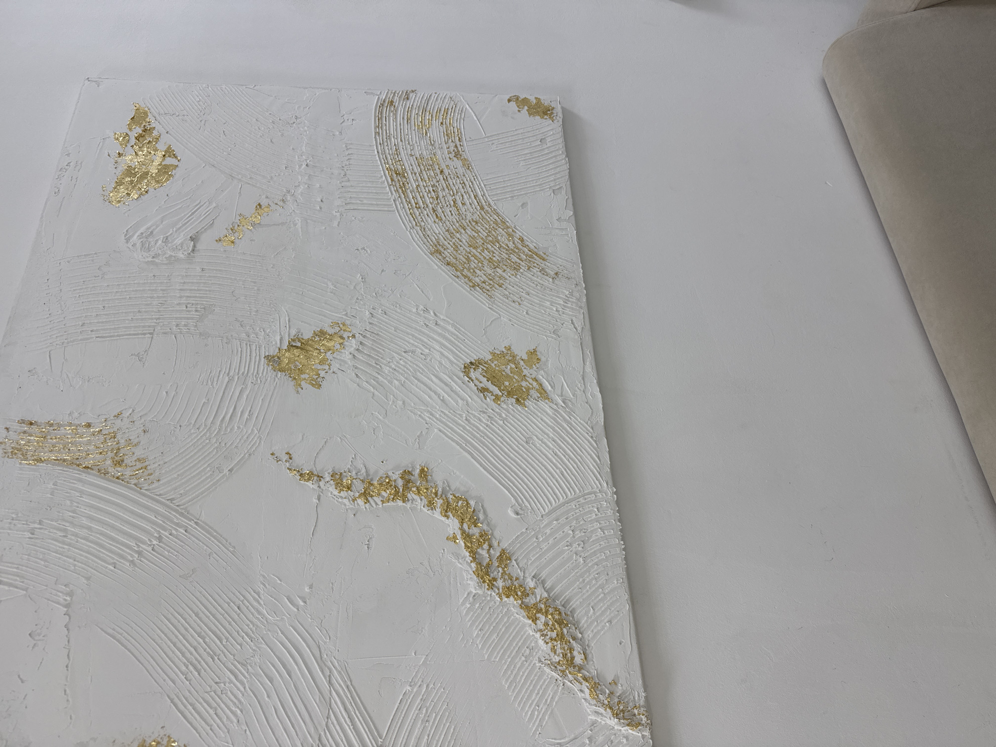 The image shows a close-up of a white textured painting with gold leaf accents, likely displayed on a white wall. Part of a light beige or off-white object, potentially furniture, is visible in the upper right corner. The location is unknown, and there is no evidence to suggest that this image was taken in the city of Chaikovsky, Russia.