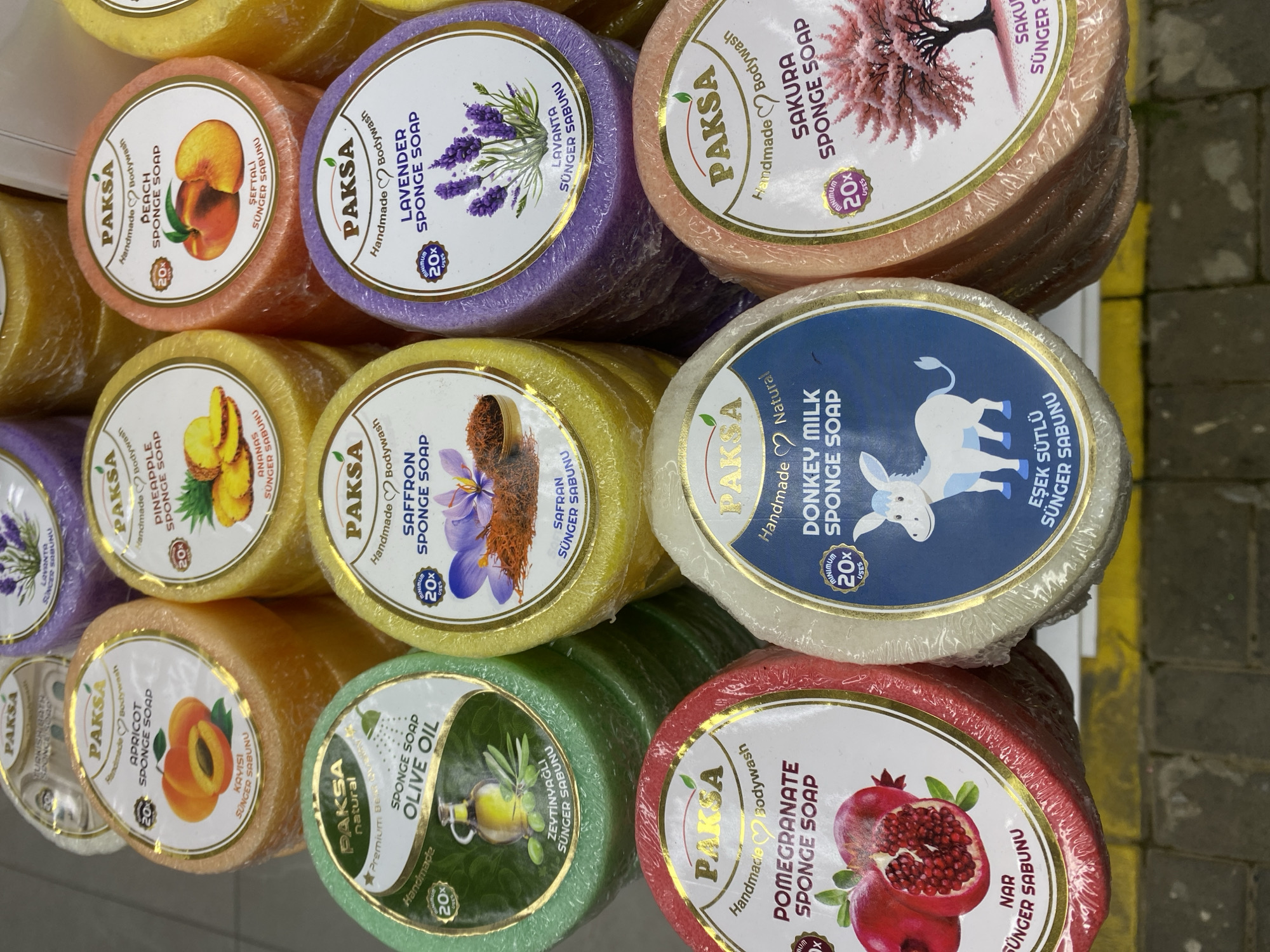 The image shows a display of round, colorful sponge soaps packaged in clear plastic wrap. The soaps are arranged in rows and columns, showcasing different varieties. Each soap has a label with the brand name "PAKSA" and the name of the soap, such as "PEACH SPONGE SOAP", "LAVENDER SPONGE SOAP", "PINEAPPLE SPONGE SOAP", "SAFFRON SPONGE SOAP", "DONKEY MILK SPONGE SOAP", "SAKURA SPONGE SOAP", "APRICOT SPONGE SOAP", "OLIVE OIL SPONGE SOAP", and "POMEGRANATE SPONGE SOAP". The labels include images of the corresponding fruits, flowers, or the donkey. The soaps are of different colors such as peach, purple, yellow, white, pink, green, and red. The image was probably taken in Alanya, Turkey, since Turkish translations are present on the labels. The location appears to be a shop or market. The soaps are neatly stacked and packaged, suggesting a commercial setting.