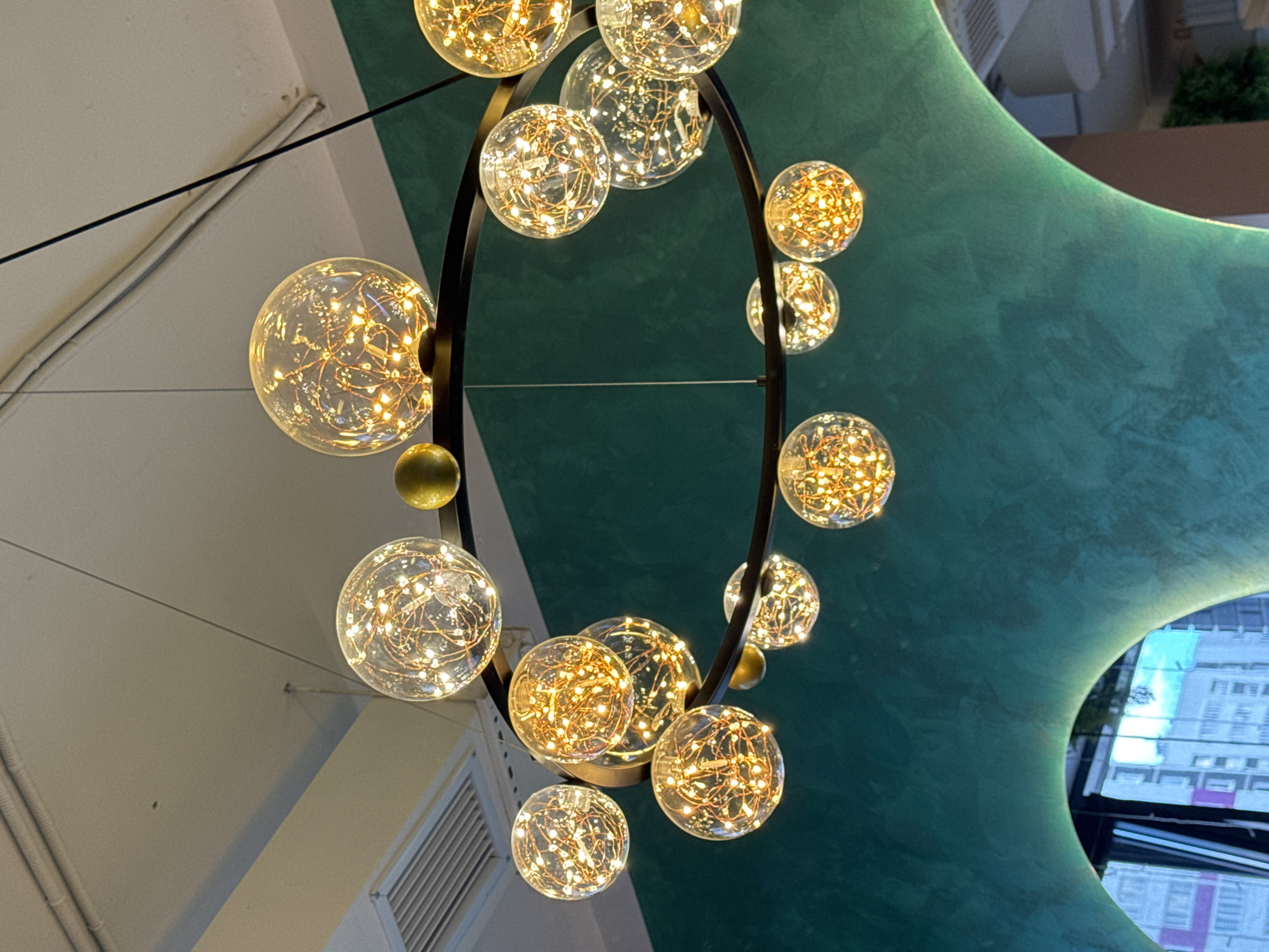 This is a medium shot, eye-level photograph of a modern chandelier with multiple glass orbs filled with fairy lights. The chandelier is suspended from thin black wires against a backdrop of a teal-colored, abstract ceiling structure and a white wall with exposed conduit.
The chandelier features a large, dark, circular frame from which numerous glass spheres hang. Each sphere contains a dense network of small, warm white LED lights that give them a glowing, festive appearance. The orbs vary in size, with some larger and others smaller, and they are arranged both within and around the circular frame.
The teal ceiling structure has a textured, organic shape, creating a contemporary and artistic feel. The white wall on the left side appears to be part of an interior space, with a visible air vent or grate. The lighting is warm and inviting, suggesting an indoor setting, possibly a restaurant, cafe, or modern living space. There is no visible text in the image. The time of day and weather are not discernible from the image.