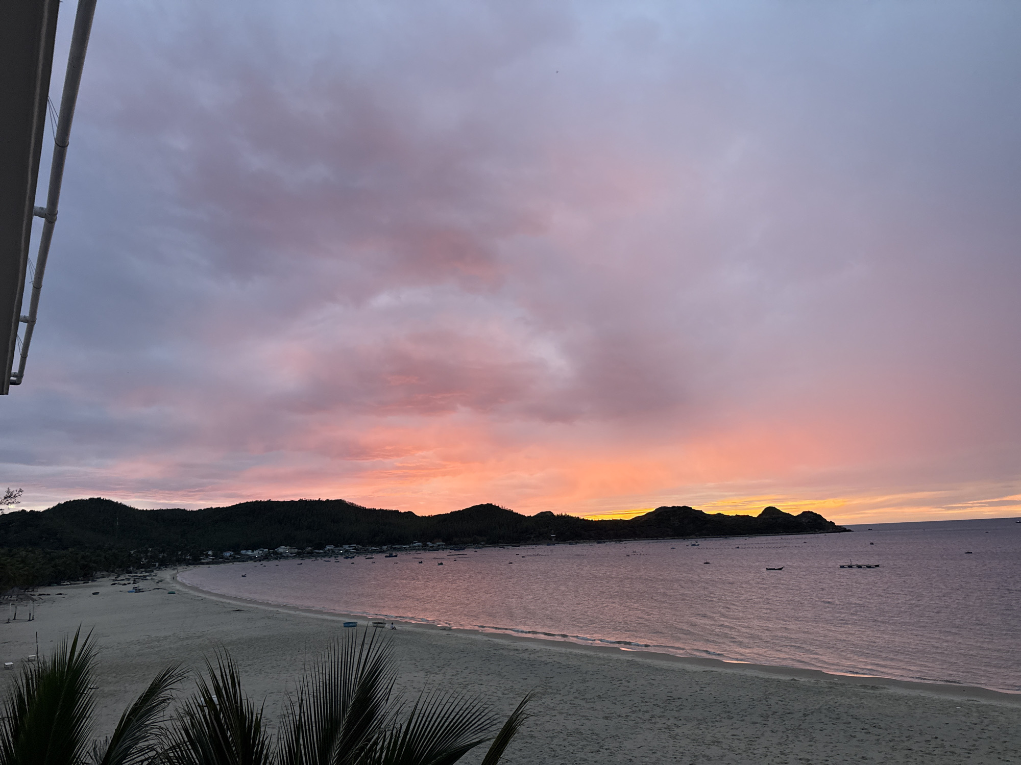 The image captures a serene sunset over a beach in Hòa Hiệp, Vietnam. A crescent-shaped beach with fine sand stretches along the coastline, dotted with a few scattered beach chairs and small boats. The calm sea reflects the vibrant colors of the sky, transitioning from a pale pink and purple near the horizon to a softer lavender higher up. Lush green hills form a backdrop to the beach, with a small village nestled along the shore, its buildings visible as white specks. Several boats are moored in the shallow waters. In the foreground, the dark silhouettes of palm fronds add a tropical touch to the scene. The sky is filled with wispy clouds, painted with hues of orange, pink, and purple, indicating the sun has recently set or is about to rise. The overall impression is one of tranquility and natural beauty.
