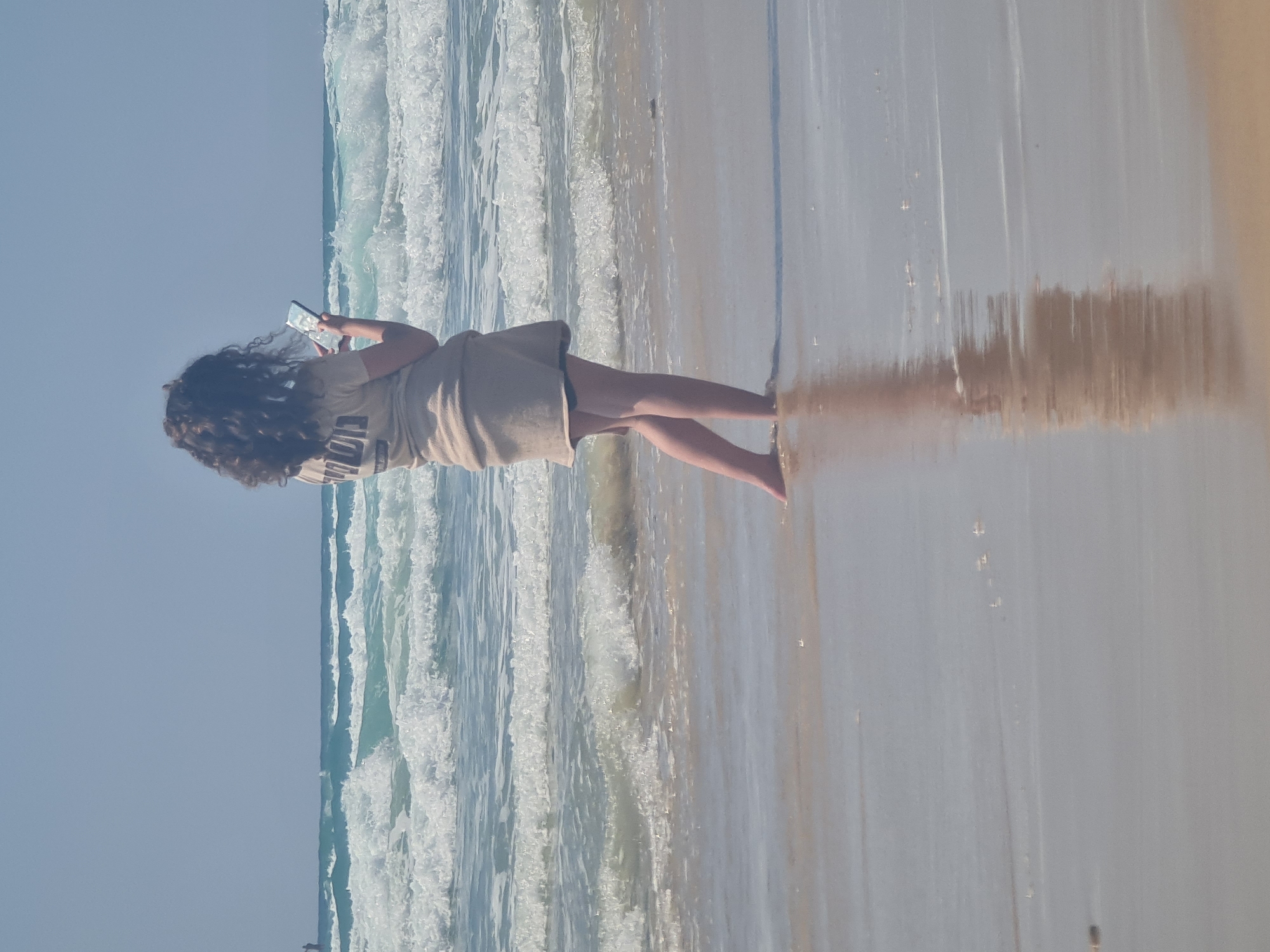A person with curly dark hair stands at the edge of the ocean, facing away from the camera. They are wearing a tan t-shirt with dark text on the back and a tan skirt. The person is holding a smartphone in their right hand, looking at its screen. Waves are breaking on the shore around their feet, and the water reflects their image on the wet sand. The sky is clear and pale blue. In the distance, a small figure can be seen standing in the water. The location is Unknown, Unknown.