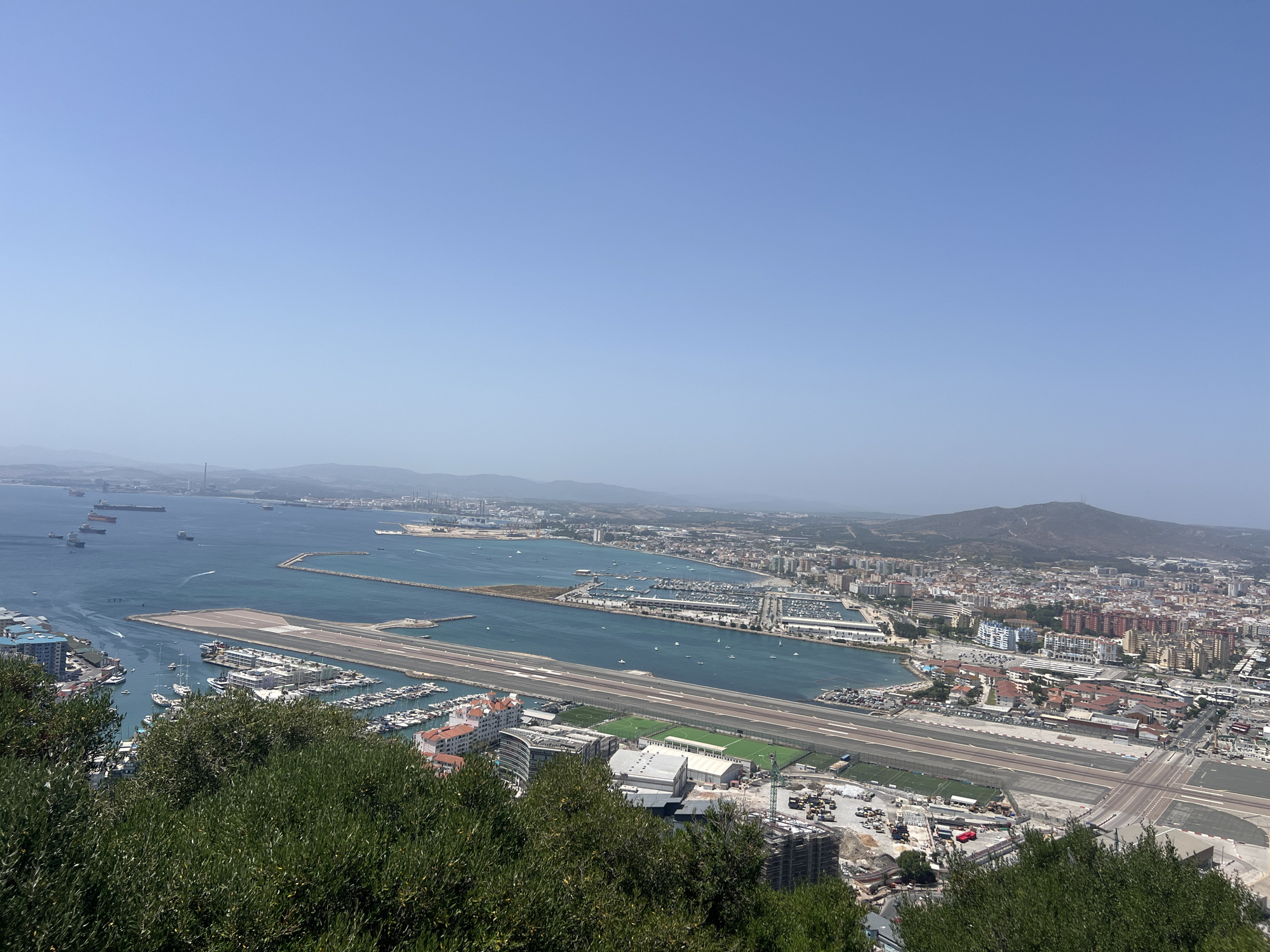 The image shows a panoramic view of the city of Gibraltar, Gibraltar, from a high vantage point. In the foreground, lush green trees and foliage partially obscure the view. Below, the scene unfolds to reveal a bustling harbor filled with numerous boats and yachts of various sizes. Beyond the harbor, a long, wide runway, characteristic of Gibraltar Airport, stretches across the landscape. The airport runway is a prominent feature, with a road crossing directly over it.
Adjacent to the runway, there are several buildings, including some with red-tiled roofs, and areas that appear to be under construction. Several sports fields, likely soccer pitches, are visible nearby. The water body is a vibrant turquoise, indicating clear conditions. In the middle ground and background, the city of Gibraltar sprawls along the coastline, characterized by a dense collection of buildings of varying heights and colors.
Further out, the bay is dotted with a multitude of ships, including large cargo vessels, indicating significant maritime activity. The distant landscape is dominated by rolling hills and mountains, shrouded in a light haze, suggesting a warm and clear day. The sky is a vast expanse of pale blue, with no clouds visible. The overall impression is one of a densely populated coastal city with a significant connection to maritime and aviation infrastructure, set against a backdrop of natural topography.
