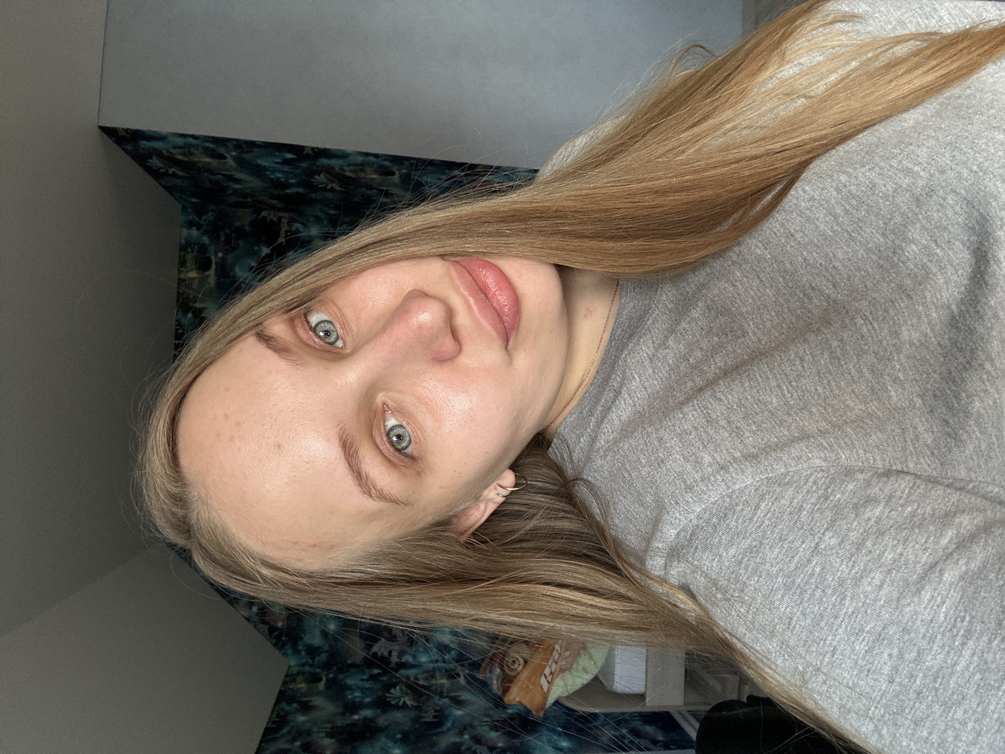 A woman with light blonde hair and blue eyes is shown in a selfie. She is wearing a grey shirt. The background consists of a wall with dark patterned wallpaper and a lighter grey wall. The location is not discernible from the image and therefore cannot be confirmed to be in Chaikovsky, Russia.