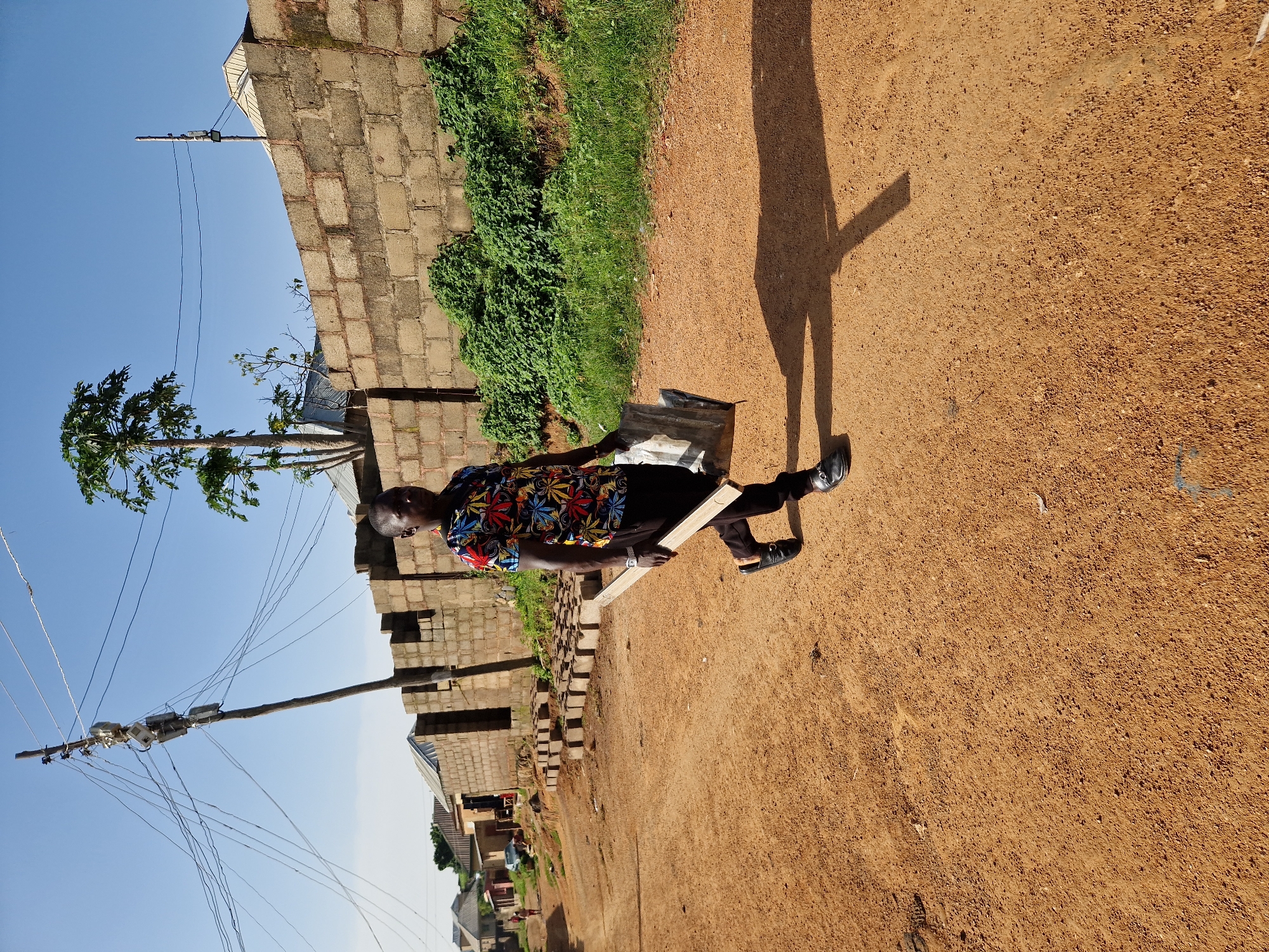 In a dusty, unpaved street in Jos, Nigeria, a man walks carrying a wooden plank and a piece of sheet metal. He is wearing a colorful, patterned short-sleeved shirt and dark trousers, with black loafers. The street is lined with brick buildings and a wall constructed from concrete blocks. In the background, a utility pole with numerous wires stands tall, and a tree with broad leaves grows near the wall. Several piles of bricks are visible along the side of the road, suggesting construction activity. The sky is clear and blue, indicating a sunny day.