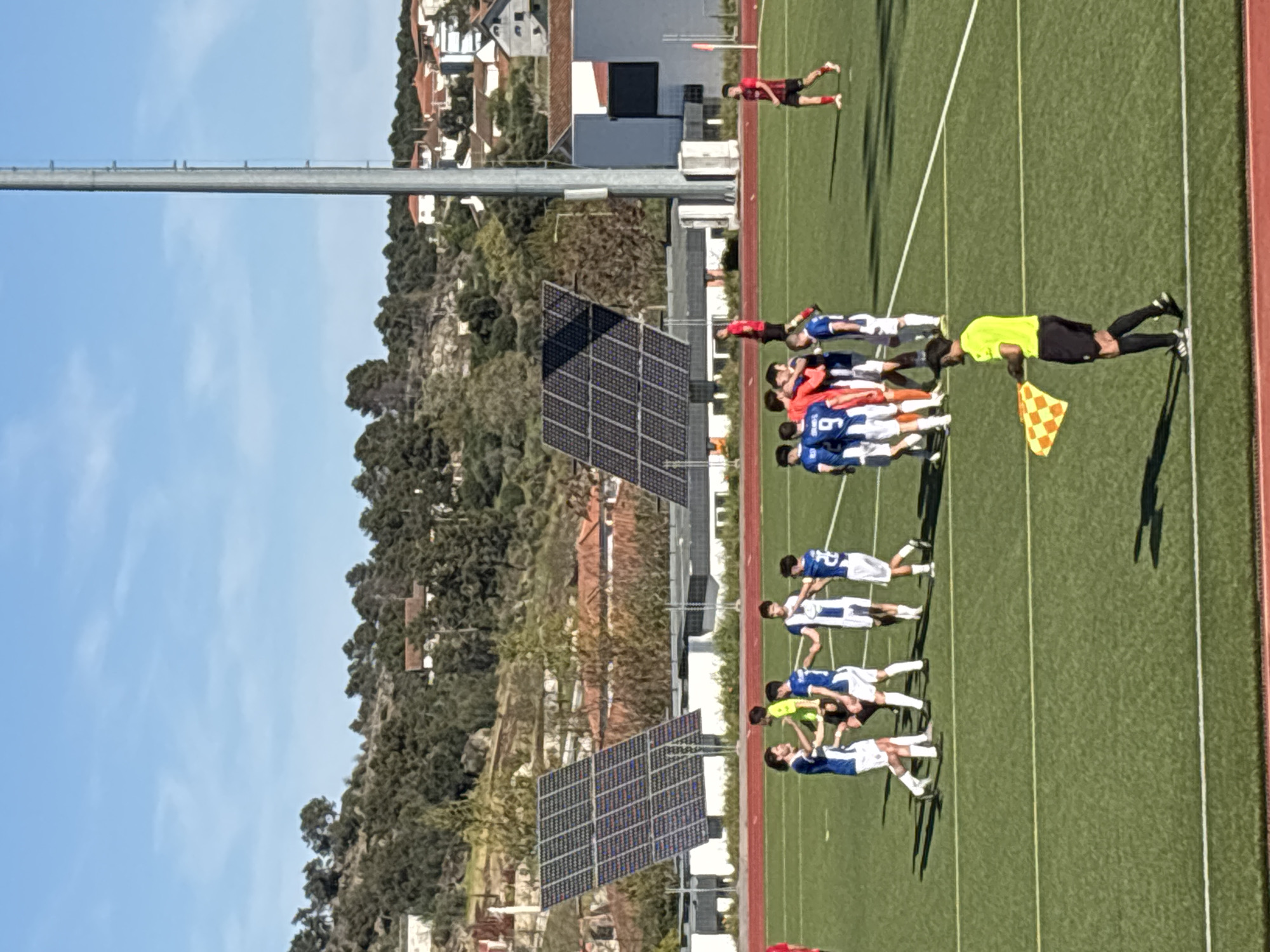 A soccer game takes place in Valpaços, Portugal. A group of players in blue and white uniforms are lined up on the green field. One man in a yellow shirt and black shorts, holding a checkered flag, stands next to the players. Another individual in red lays on the field in the background. Solar panels stand to the side of the field. Buildings and trees are visible in the background under a clear blue sky.