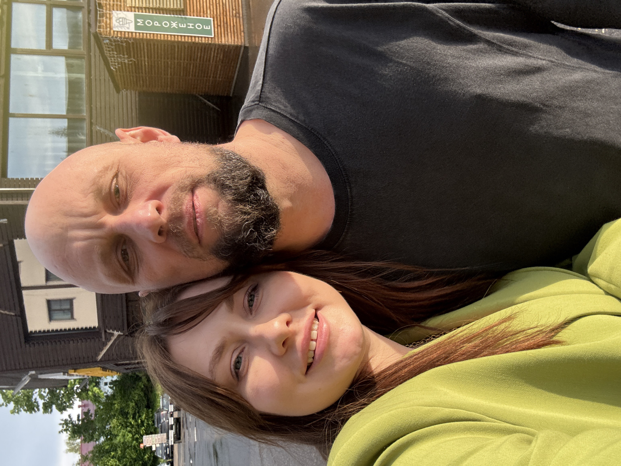 A medium close-up shot captures a man and woman outdoors. The man is bald with a dark beard and is wearing a black shirt. The woman has long brown hair and is wearing a light green shirt. The image is oriented upside-down, relative to typical orientation conventions. Behind the couple, out-of-focus buildings and trees can be seen. In the top left corner of the frame, a building with a sign in Russian that says "мороженое" (ice cream) is visible. The scene is located in the city of Peresvet, Russia.