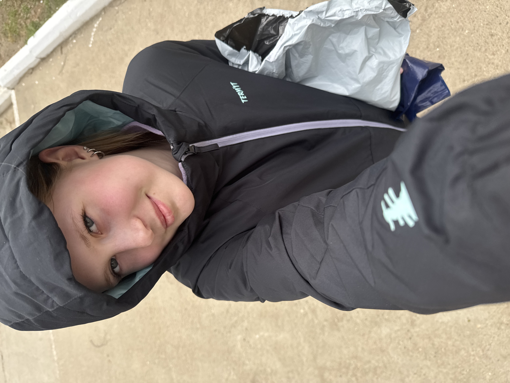 A young person, possibly female, is captured in a high-angle selfie, looking up at the camera. The image is rotated, showing the person's head on the left, tilted slightly, and their body extending towards the right. They have light brown hair peeking from under a dark grey or black hooded jacket with a light blue or mint green lining visible around the hood. The person has light-colored eyes and a subtle, friendly smile on their face. Their left ear is visible, adorned with multiple silver hoop earrings and a stud.
The jacket features a light purple zipper and the brand name "TERMIT" in a light blue or mint green font on the upper right arm/shoulder area. Another logo, abstract and also in light blue or mint green, is visible on the lower right sleeve. In the background, behind the person's right shoulder, there are what appear to be two packages or bags – one grey and crumpled, the other dark blue.
The setting appears to be outdoors during the daytime, possibly overcast, given the soft lighting. The ground is a light, sandy color, possibly concrete or asphalt, with a lighter-colored curb or edge visible in the upper left. The overall impression is a casual outdoor moment. The location context is Chaikovsky, Russia.