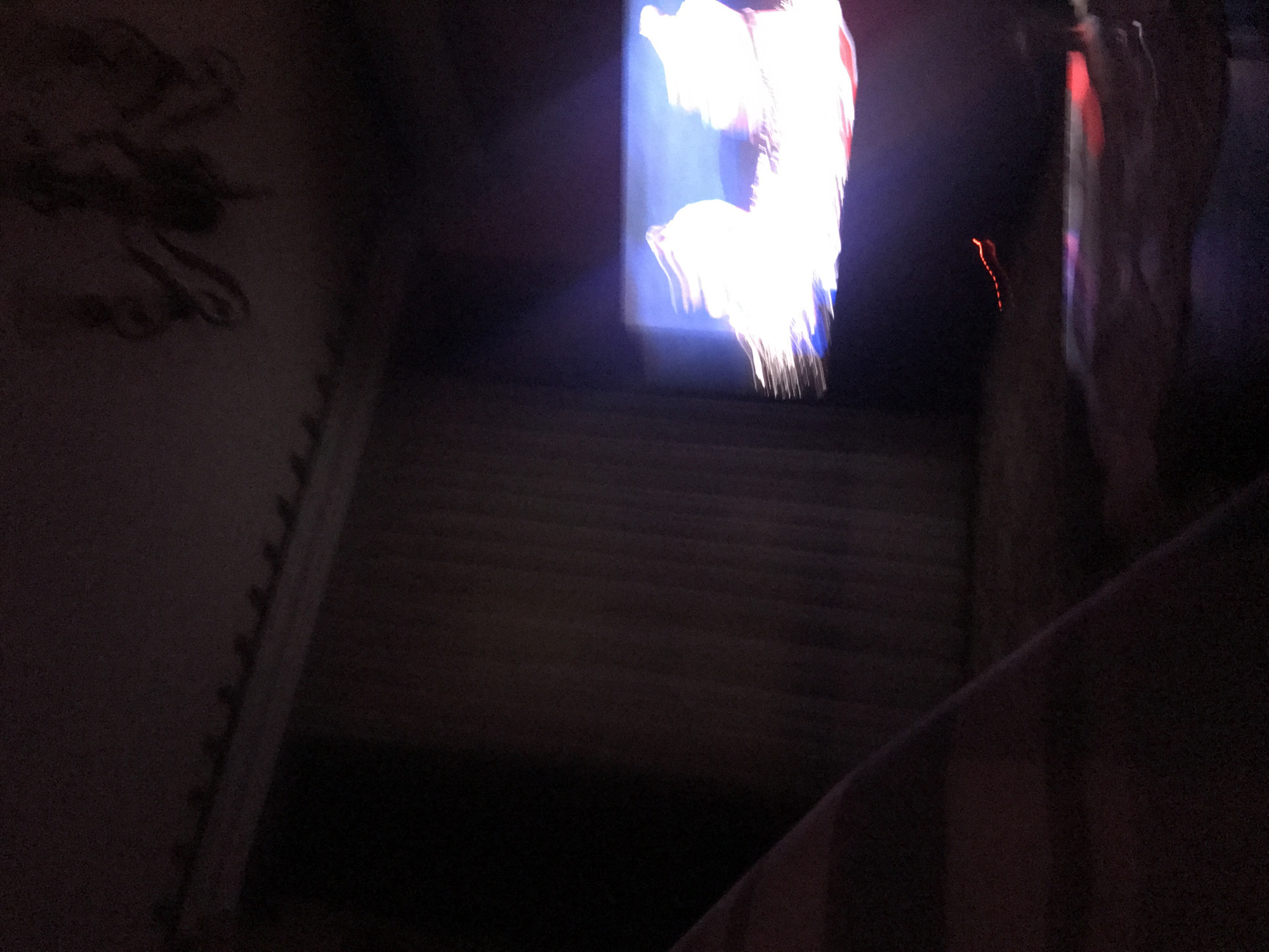 The photograph appears to show a darkened indoor space, possibly a stairwell or a narrow hallway, illuminated by an unseen light source. A bright, indistinct image is visible in the upper center, possibly from a window or a screen, while the lower half of the image is obscured by darkness. The presence of wall decorations on the left and the steps suggests a built environment. Yerevan, Armenia is not discernible from the presented image.