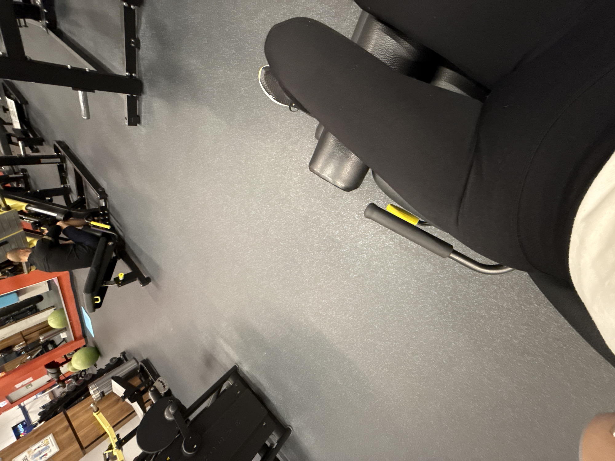 The image displays an indoor gym environment, captured from a first-person perspective, looking down and forward. The viewpoint originates from a person seated on a gym machine, whose legs, clad in black leggings and white athletic shoes with white socks, are visible in the foreground on the right side of the frame. Part of a white top is also visible, along with the black padded seat and a yellow adjustment lever of the machine.
The floor is a prominent feature, showing a light gray, speckled texture typical of a gym. A light gray wall runs across the upper portion of the scene.
In the mid-ground and background on the left, another individual is seen actively using a different piece of black gym equipment. This person has short, light-colored hair and is wearing a dark top and dark lower garments. Their face is indistinct. Surrounding this person are various other black gym machines, including racks and benches, indicating a well-equipped fitness facility. A large mirror with a red frame reflects parts of the gym, including some green stability balls, and a wooden cabinet or shelf with a small, unreadable sign is also visible on the far left.
The scene depicts a typical gym setting where individuals are engaged in exercise. The lighting is consistent with an indoor commercial establishment. There are no discernible emotional expressions, and no explicit geographical cues within the image itself, though the context indicates it could be a gym in Sofia, Bulgaria. No legible text is present.