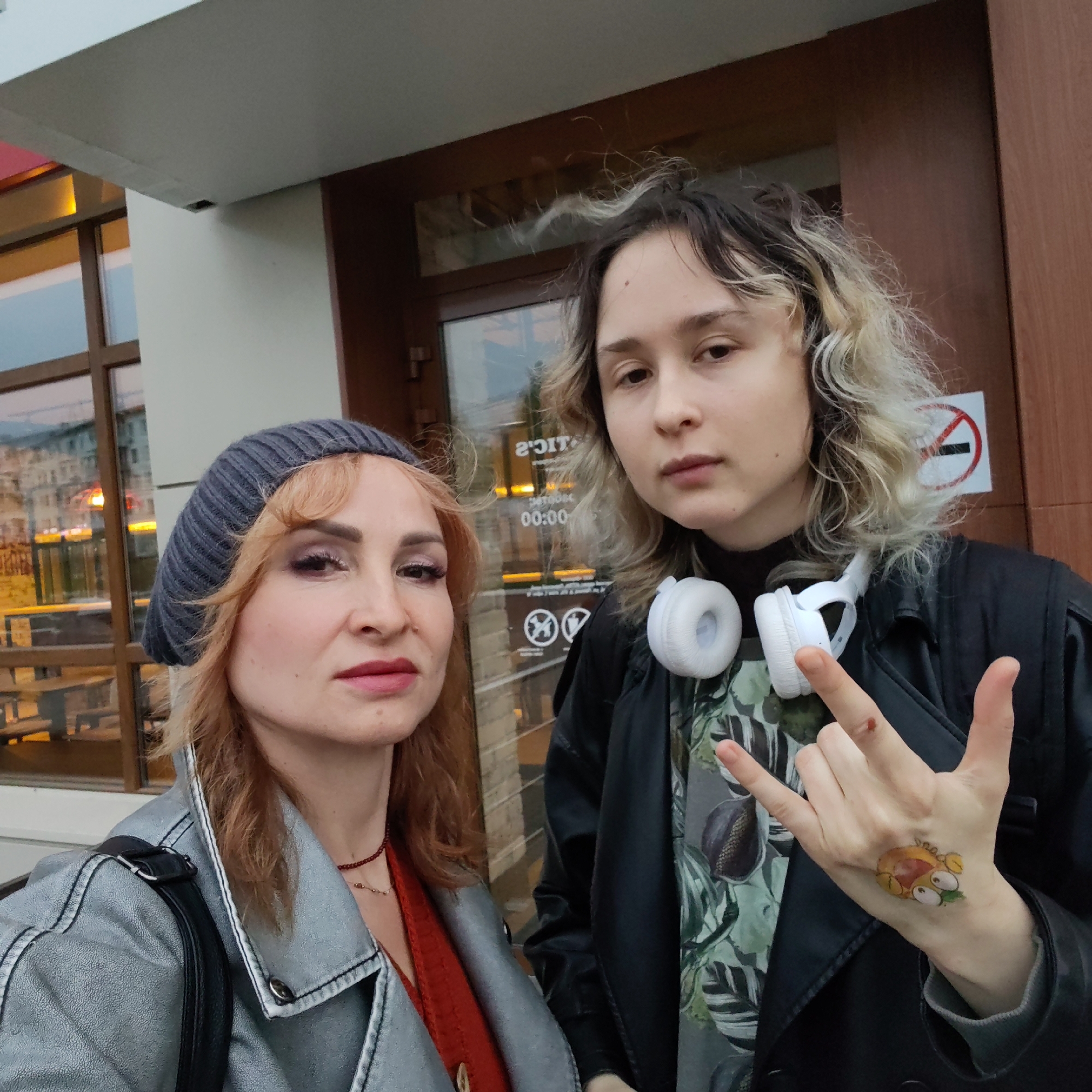 Two women are standing in front of a building with a glass door in the city of Chaikovsky, Russia. The woman on the left wears a gray knit hat, a metallic silver jacket, and an orange top, with a black shoulder bag. The woman on the right wears a black leather jacket over a patterned shirt, with white headphones around her neck, and gives a "rock on" hand gesture with a cartoon tattoo on her hand. A "no smoking" sign is visible on the door behind them. The glass door shows reflections of the surrounding urban environment.