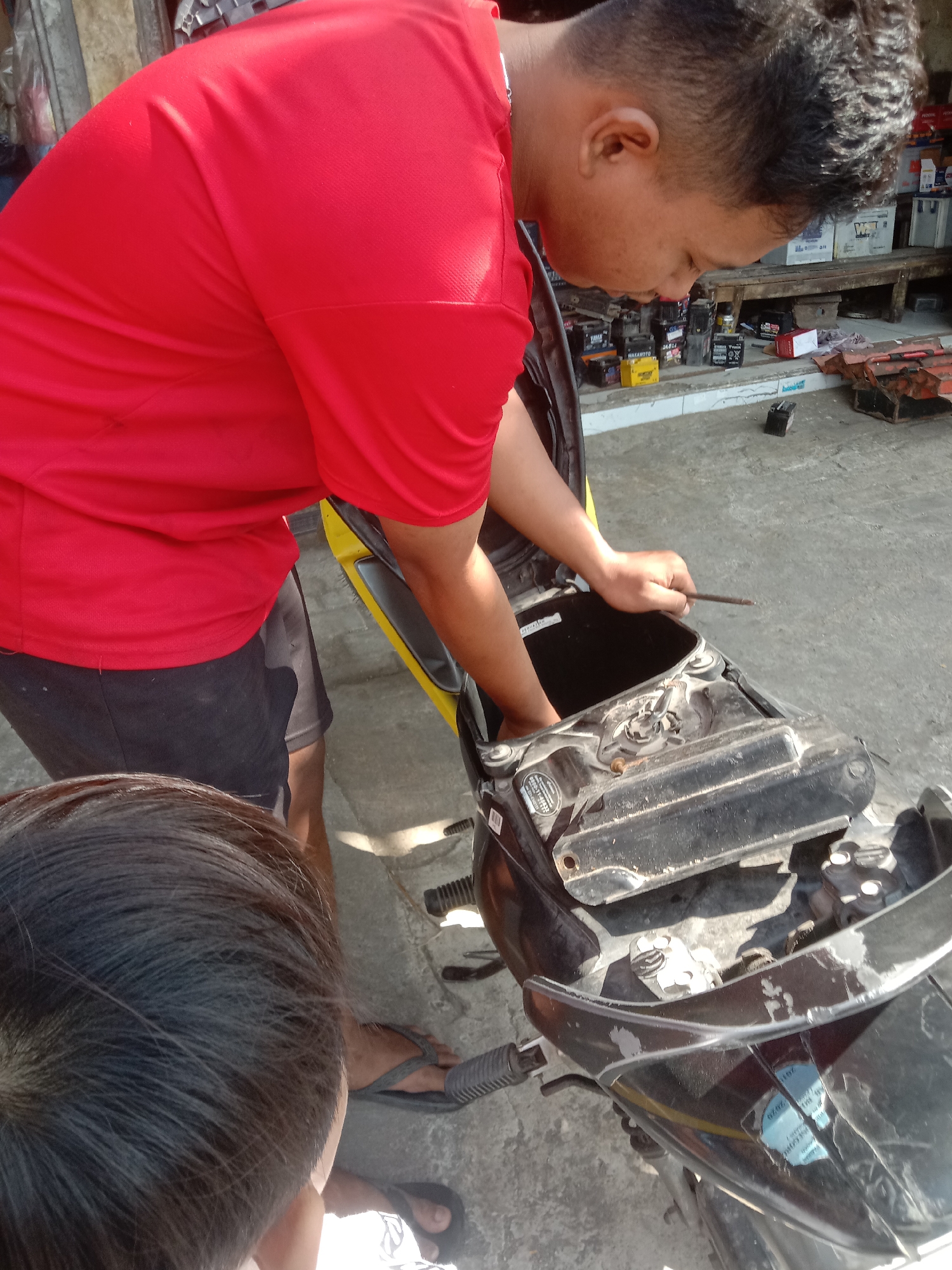 In Pasinan, Indonesia, a person wearing a red t-shirt and black shorts is working on a motorcycle. The person is hunched over, with their right hand inside the motorcycle's storage compartment, holding a long, thin tool. Another person's head, seen from behind, is in the foreground, suggesting they are observing or assisting. The scene appears to be outdoors, with a concrete ground and a background that includes shelves filled with various items, possibly in a shop or workshop setting. The lighting suggests it is daytime, with sunlight casting shadows.