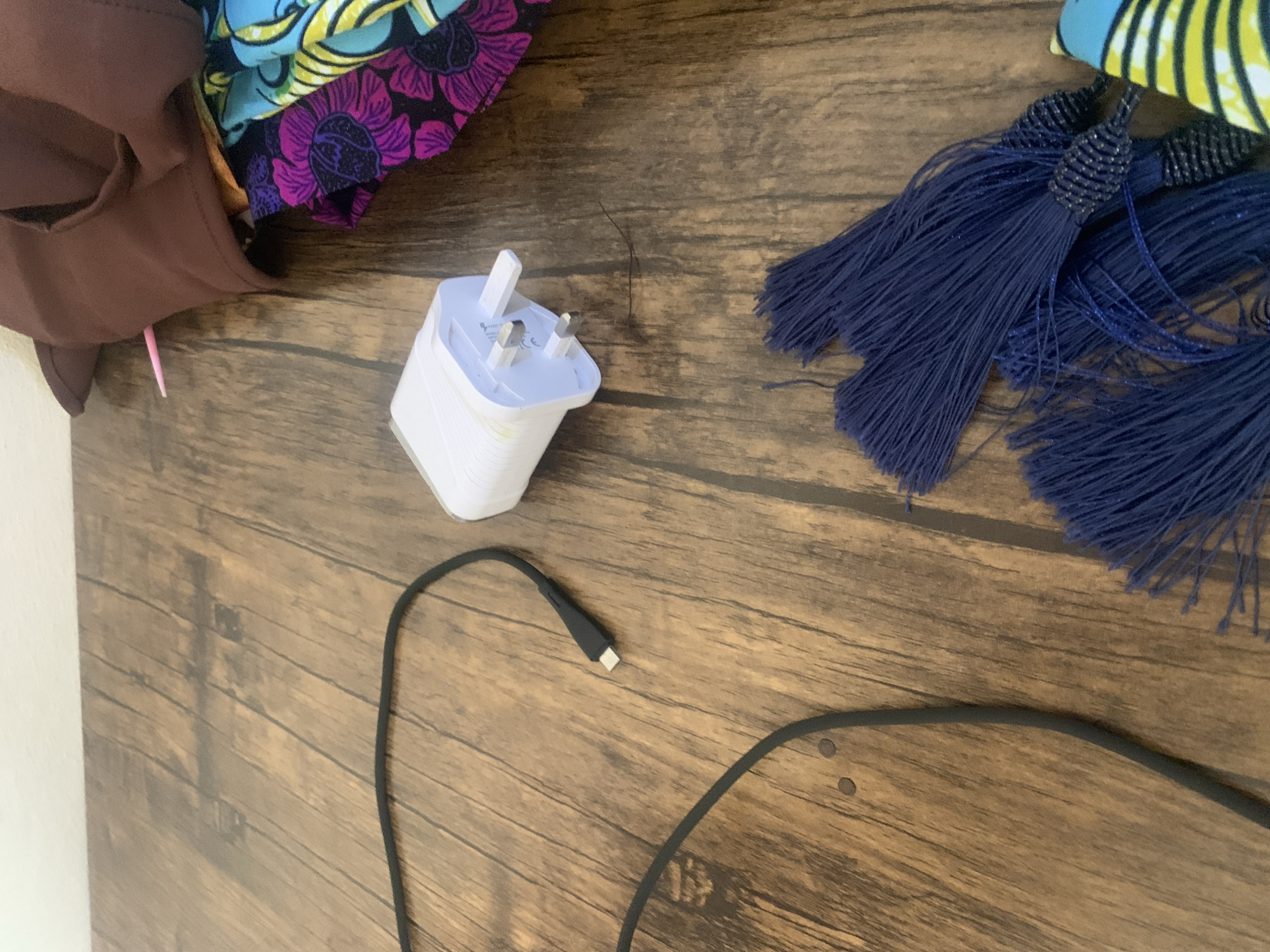 A white wall is visible at the top of the image, bordering a wooden surface. The wooden surface is textured, with visible grain patterns and variations in color, suggesting it may be a table or floor.
In the middle of the frame, a white USB charger is placed on the wooden surface. To the left of the charger, a black USB cable is visible, with its connector end pointing towards the charger.
In the upper right corner, a piece of brown fabric is draped over what appears to be colorful patterned fabric, possibly clothing or a textile. A pink sewing pin is stuck into the brown fabric.
In the lower right portion of the image, several dark blue tassels are arranged on the wooden surface. They are bunched together, with some individual strands of thread visible. Part of a brightly colored patterned fabric is visible next to the tassels.
The image appears to be a still life, focusing on the arrangement of these objects on a wooden surface, with a plain white wall in the background. There are no people or animals in the image, and no actions or interactions are depicted. The lighting is moderate, casting some soft shadows. The overall impression is one of domestic clutter or a workspace.
The image does not contain any identifiable landmarks or features that would allow for its precise location within Jauro Yinu, Nigeria.