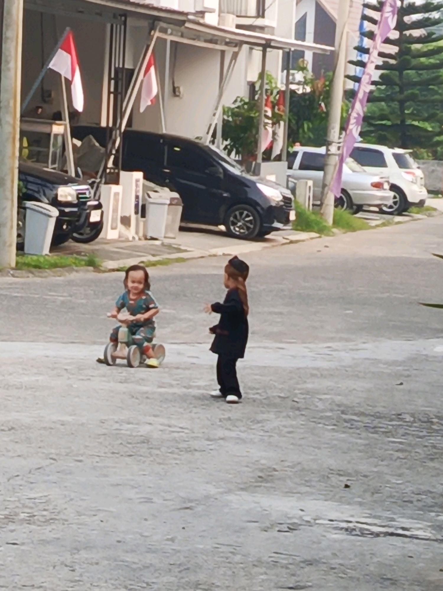 In the city of Ciomas, Indonesia, two young children are depicted outdoors in what appears to be a residential street. The child on the left is actively riding a small, three-wheeled toy bicycle, wearing a patterned outfit and yellow shoes. Their gaze is directed forward. The child on the right is standing and facing away from the viewer, dressed in dark clothing and a small hat, with their right hand raised. Behind the children, two Indonesian flags are prominently displayed on poles near a building with an overhang. Several vehicles are parked in the background, including a black car on the left and a silver SUV to the right. The environment suggests a typical street scene with buildings and paved surfaces, possibly a quiet residential area.