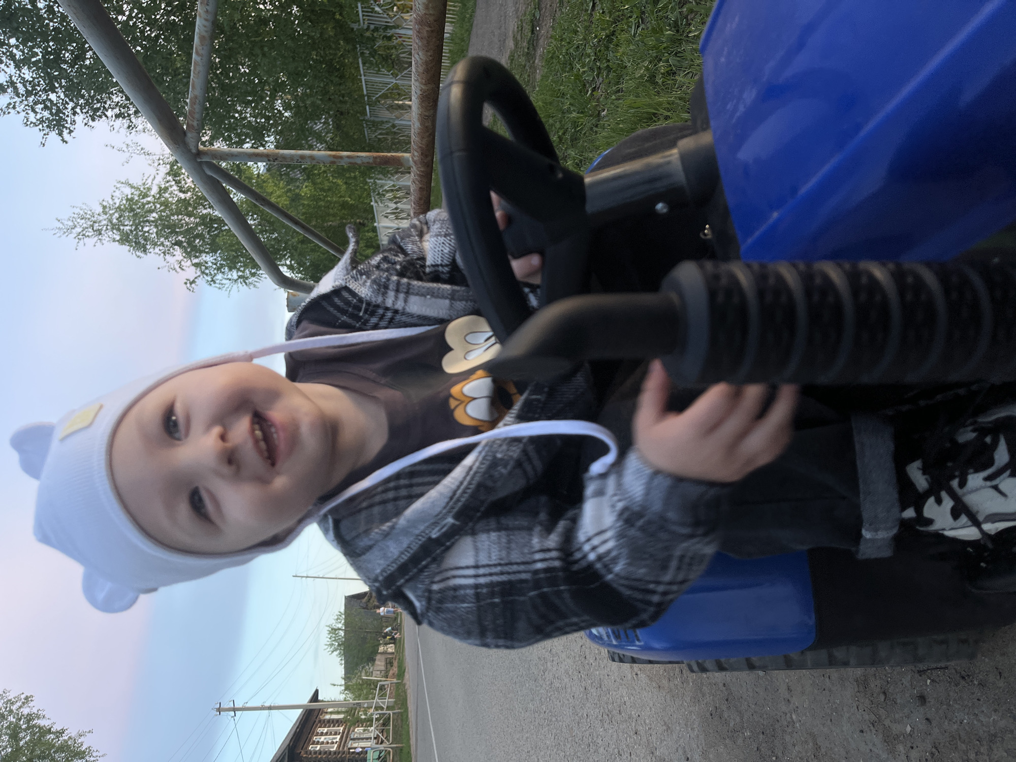 A toddler wearing a white hat with bear ears and a gray and black plaid jacket is sitting on a blue toy tractor, holding the black steering wheel. The location is the city of Фоки, Russia, as evidenced by the wooden houses in the background along a paved road. Overhead power lines are visible. The environment appears to be dusk.