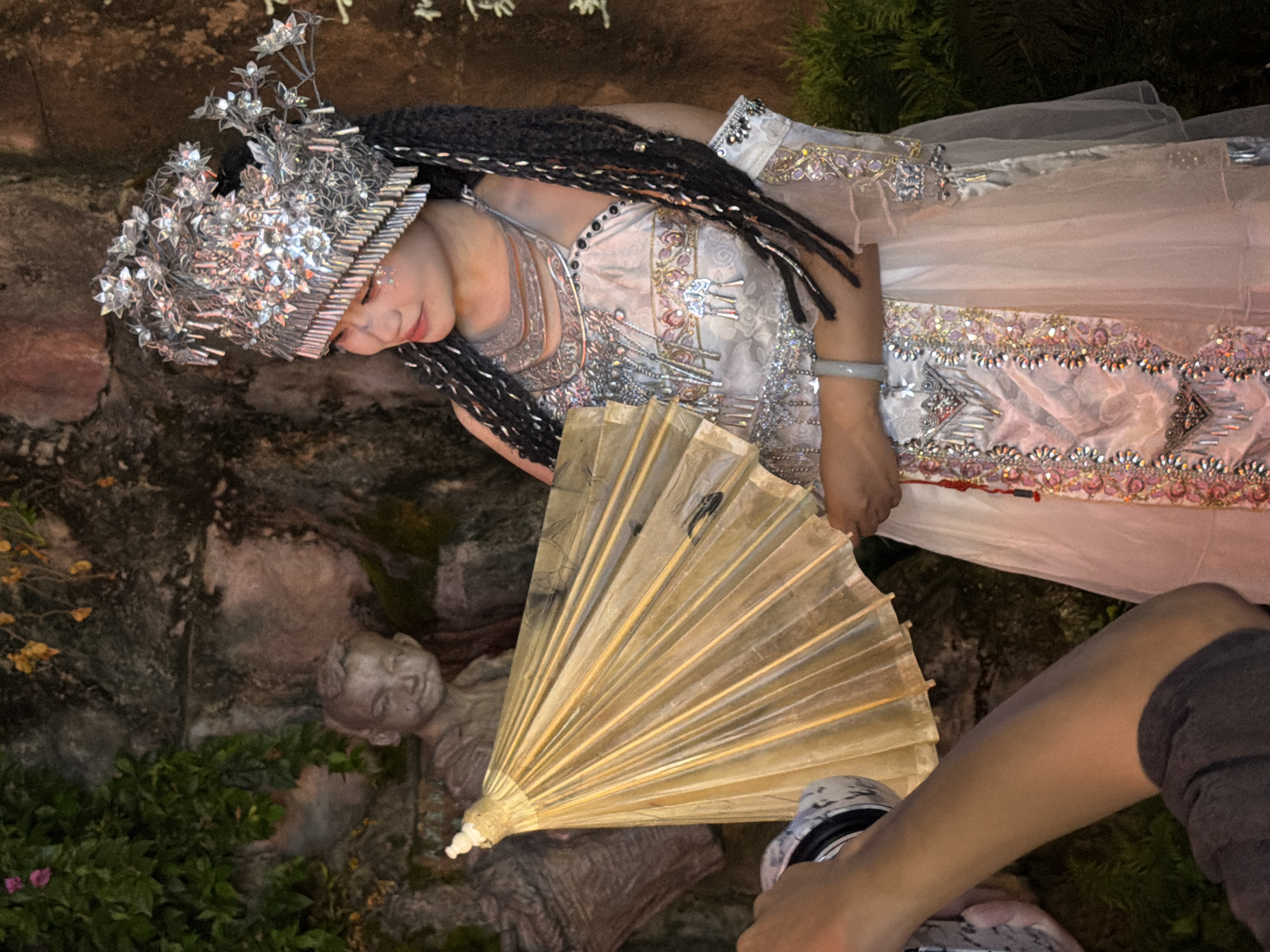 This media file features a person dressed in an elaborate, ornate costume, possibly for a cultural performance or festival. The costume is richly decorated with intricate beadwork, sequins, and metallic embellishments. The person wears a striking headdress adorned with numerous silver-toned floral and geometric elements, and their hair is styled in long, dark braids. They also appear to be wearing a delicate, light-colored veil. In front of the person, a golden fan is held open, its bamboo ribs fanning out. The scene appears to be outdoors, with a stone wall and lush greenery in the background. A small stone sculpture of a child's head is visible among the foliage. The lighting suggests it may be late afternoon or early evening, casting a warm glow on the scene. The overall impression is one of theatricality and cultural display. The location context provided is Yuzhong District, China. There is no visible text in the image.