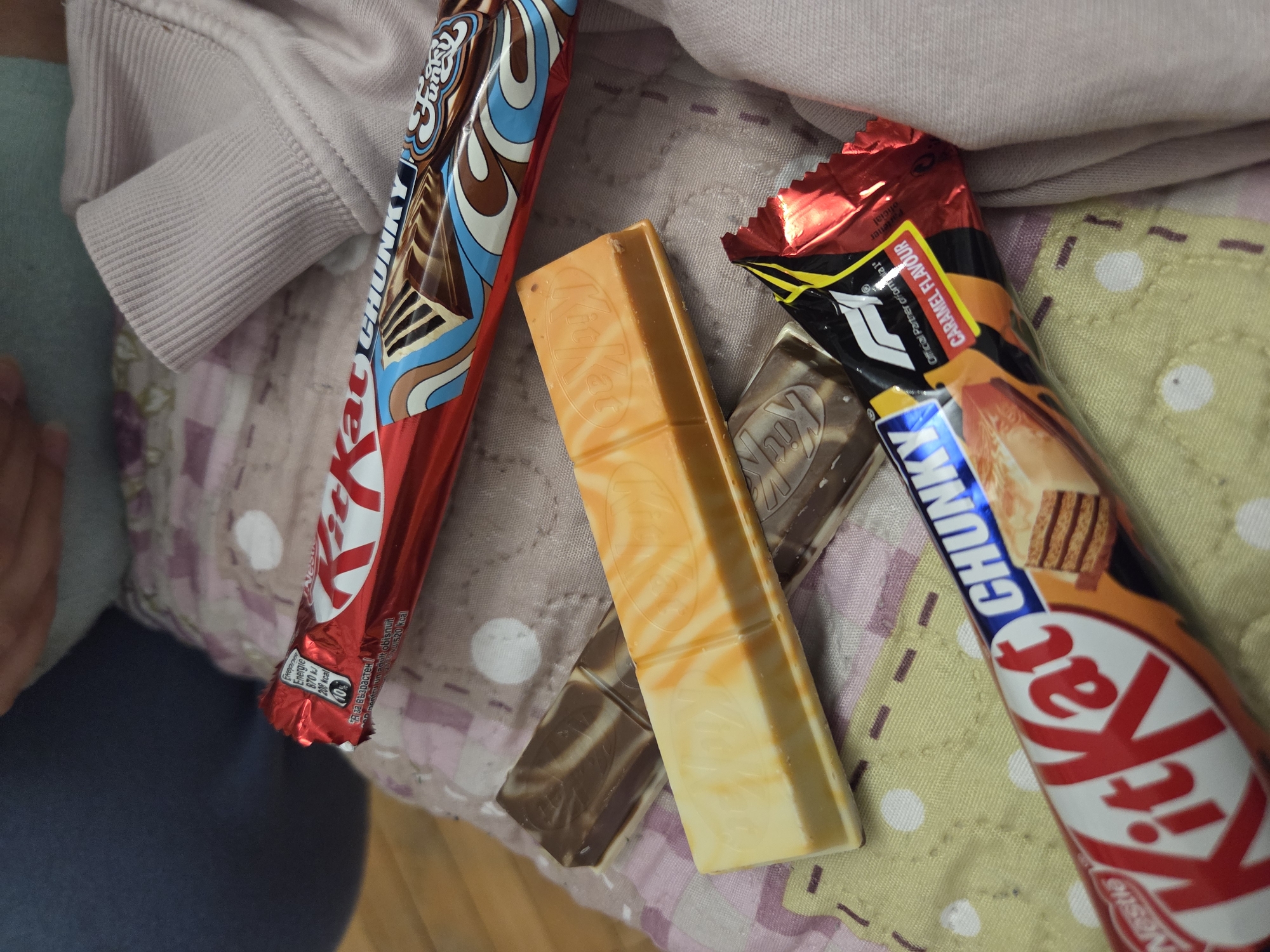 The image shows several unwrapped Kit Kat Chunky chocolate bars on what appears to be a bed or a padded surface. There are three distinct flavors visible: a "Funky" flavor in a red and blue wrapper, a "Caramel Flavour" in an orange and red wrapper, and two unwrapped chocolate bars with marbled patterns (orange-white and brown-white). The wrappers are partially torn, indicating they have been opened. The scene is casually arranged, suggesting a personal setting. There is no information to place the image in Pleven, Bulgaria.