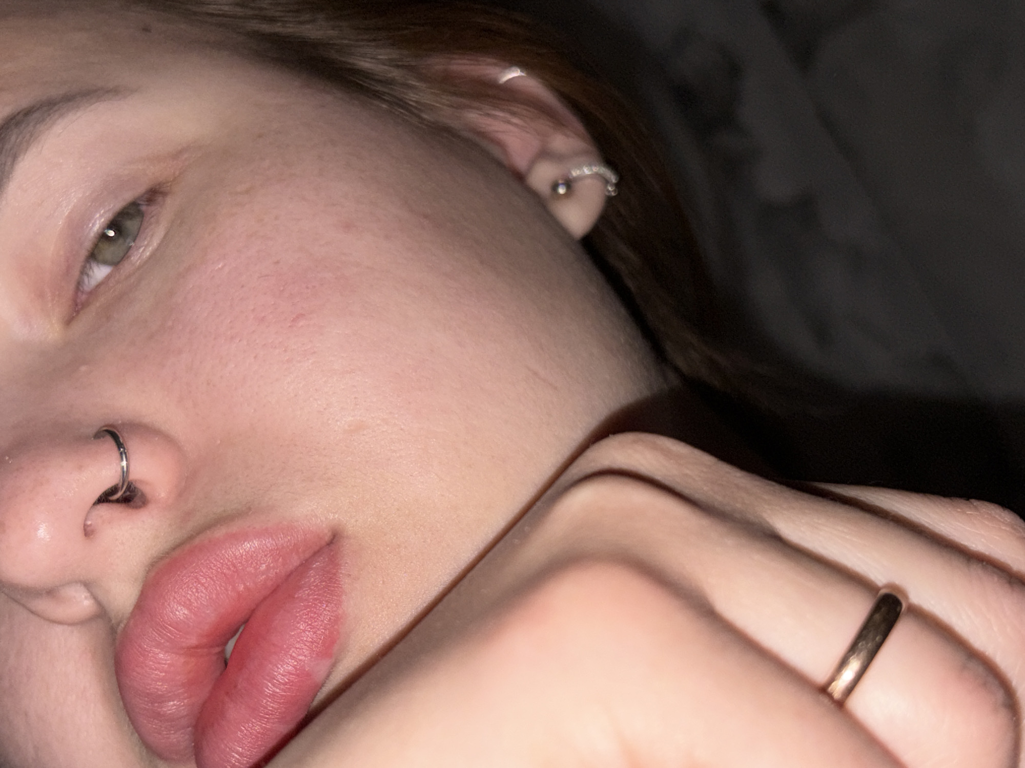 A close-up, intimate portrait captures a person reclining, with their face and a hand prominently featured. The left side of the person's face is visible, including their left eye, nose, and mouth. The eye, a striking shade of green or hazel, looks directly towards the viewer. The person has a silver septum piercing in their nose and at least two silver hoop earrings in their left ear. Their lips are full and slightly parted, showing a natural pinkish-red hue and some texture.
A hand is positioned near the lower part of the face, with fingers partially obscuring the chin and cheek area. A simple, thin gold-toned band ring is worn on one of the fingers. The skin appears natural, showing pores and some minor redness on the cheek area. Dark brown hair frames the face and is visible near the ear.
The lighting is soft and subdued, suggesting an indoor setting, possibly during the evening or night. The background is largely out of focus and dark, hinting at a soft surface like bedding or a dark fabric. The overall mood is relaxed and quiet. No text is visible in the image.