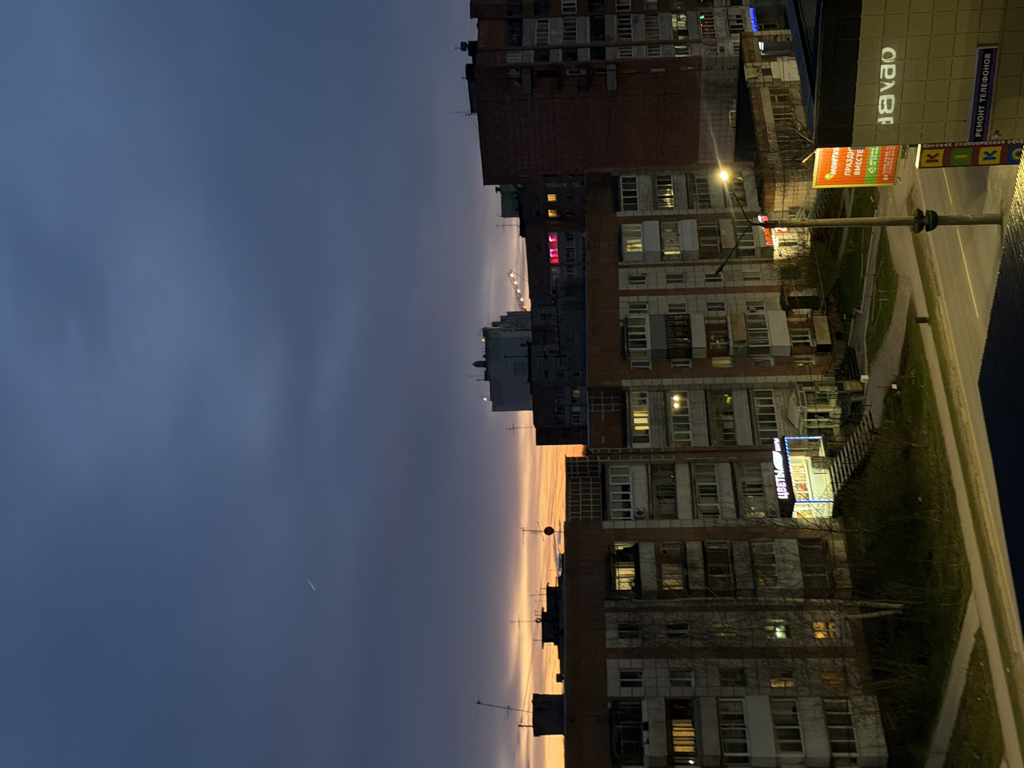 The image depicts a dusk scene in Perm, Russia, with apartment buildings lining a street. The sky is a gradient of deep blue to orange near the horizon. Buildings are multi-story, with lit windows. Street lights illuminate the pavement, and advertisements are visible on buildings. There are no visible people in the image.