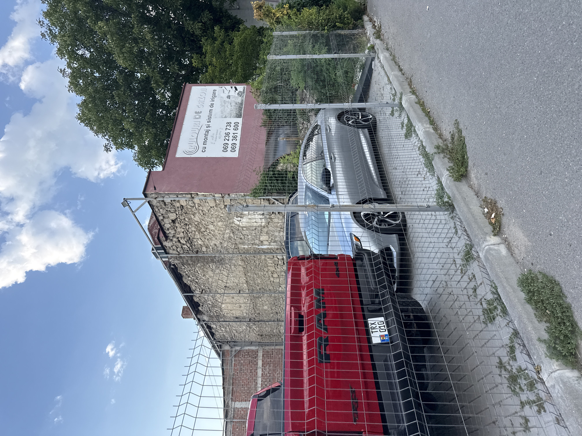 The image captures an outdoor scene in Chișinău, Moldova. In the foreground, a metal chain-link fence runs horizontally across the frame. Behind the fence, a red pickup truck with "RAM" branding is parked, followed by a silver sedan. The ground surface consists of interlocking paving stones. To the right of the vehicles, a section of a building with a dark red facade is visible, featuring a white rectangular sign with text and graphics. The sign appears to be an advertisement, possibly for lawn care services, with Romanian text that translates to "Lawns with installation and irrigation system." Two phone numbers are also displayed. Adjacent to the building and behind the fence, a stone wall with visible brickwork is present. Green trees and foliage are seen in the background. The sky above is blue with scattered white clouds. A small patch of weeds is growing along the concrete curb separating the paved area from the asphalt road in the lower right corner of the image. There are no people visible in the scene.