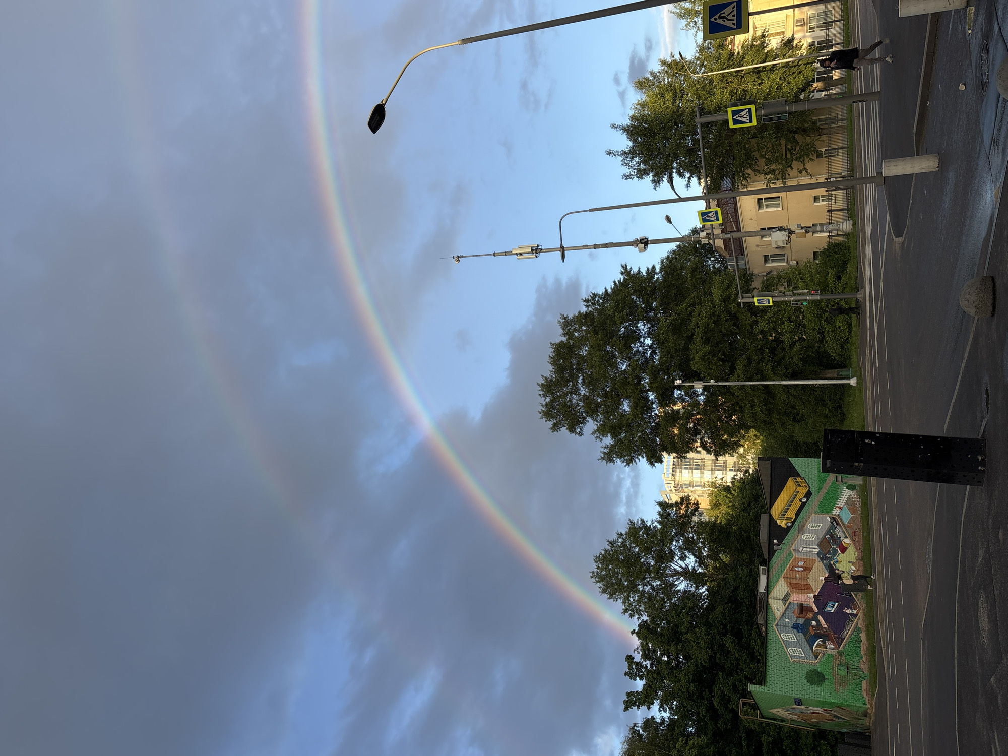 The image shows a cityscape featuring a double rainbow. In the foreground, there's a street with a mural on a building, depicting houses and a yellow school bus. Overhead pedestrian crossings with yellow signs are visible, indicating a crosswalk. Streetlights are present. The rainbow arches across the sky, partially obscured by trees. In the background, buildings are visible. The location is Moscow, Russia. A person is walking on the crosswalk.